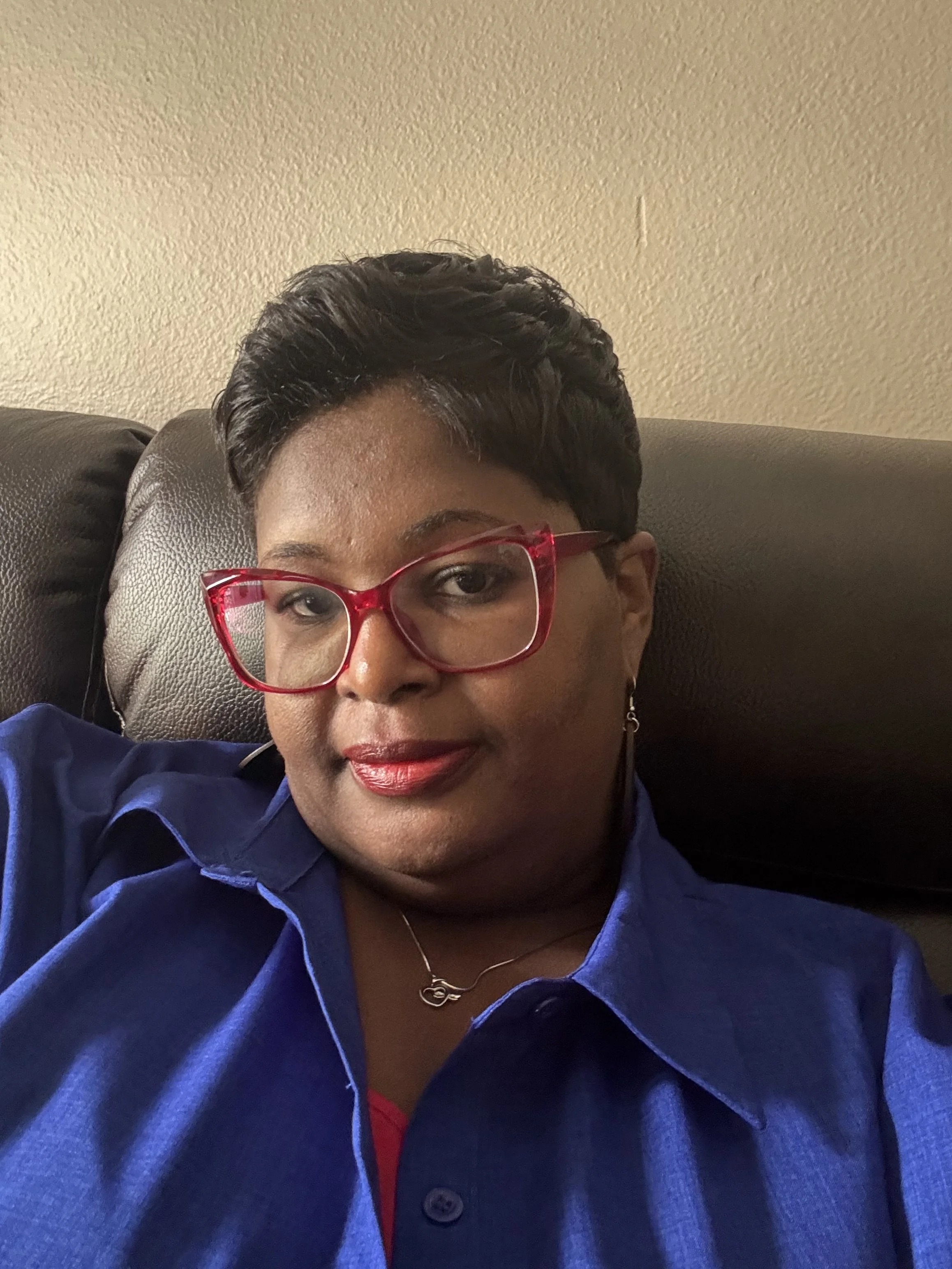 A woman with short black hair wearing red glasses, a blue shirt, and a silver necklace, sitting on a dark brown couch against a beige textured wall.