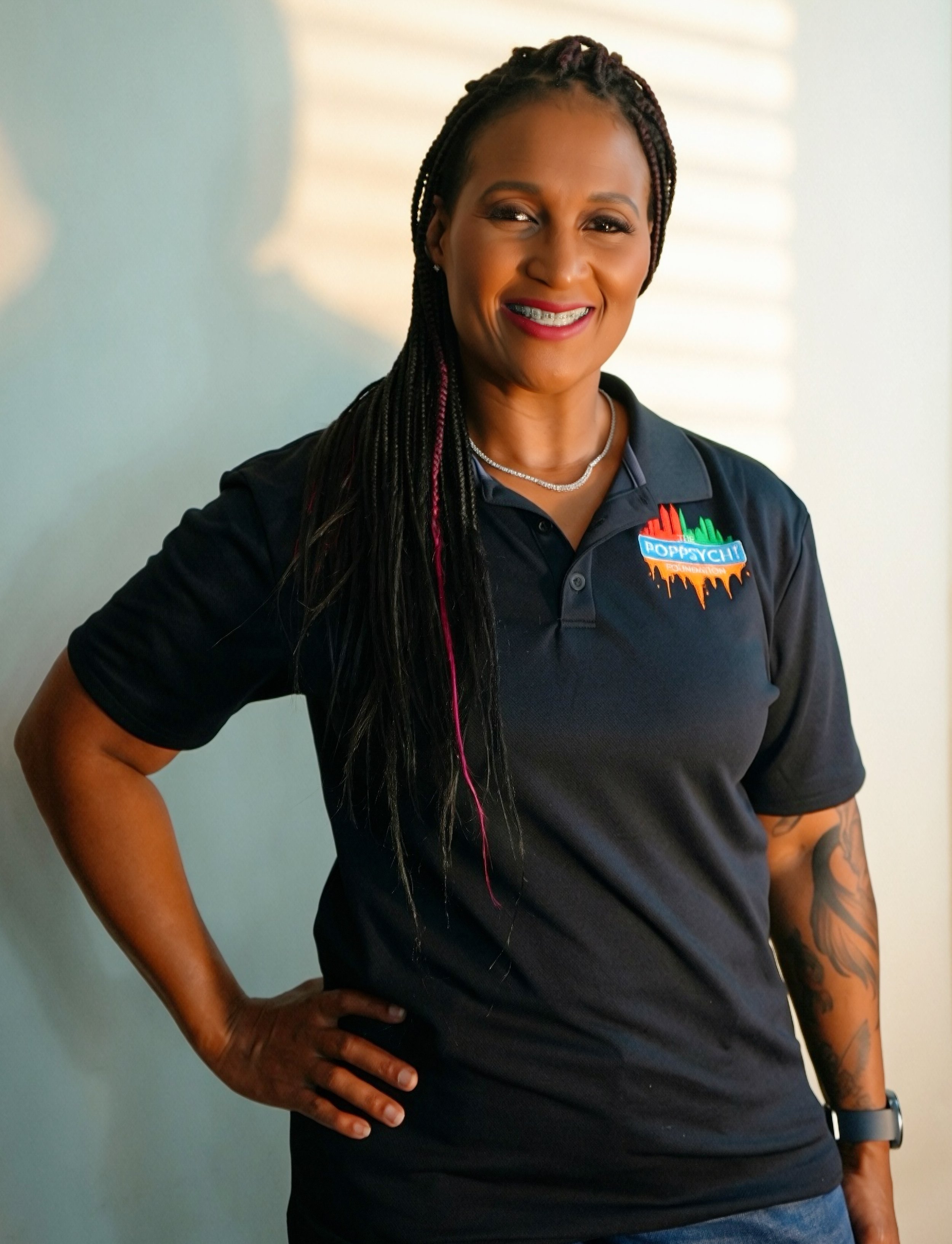 A woman with braided hair, braces, and a necklace, smiling and wearing a black polo shirt with a logo that says 'Poppy CHI Foundation'.