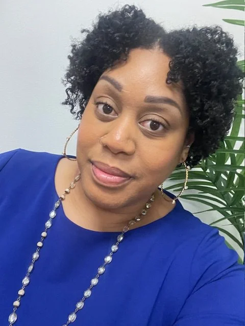 A woman with short, curly black hair, wearing a blue top, earrings, and a beaded necklace, smiling at the camera with a white background and green plant in the background.