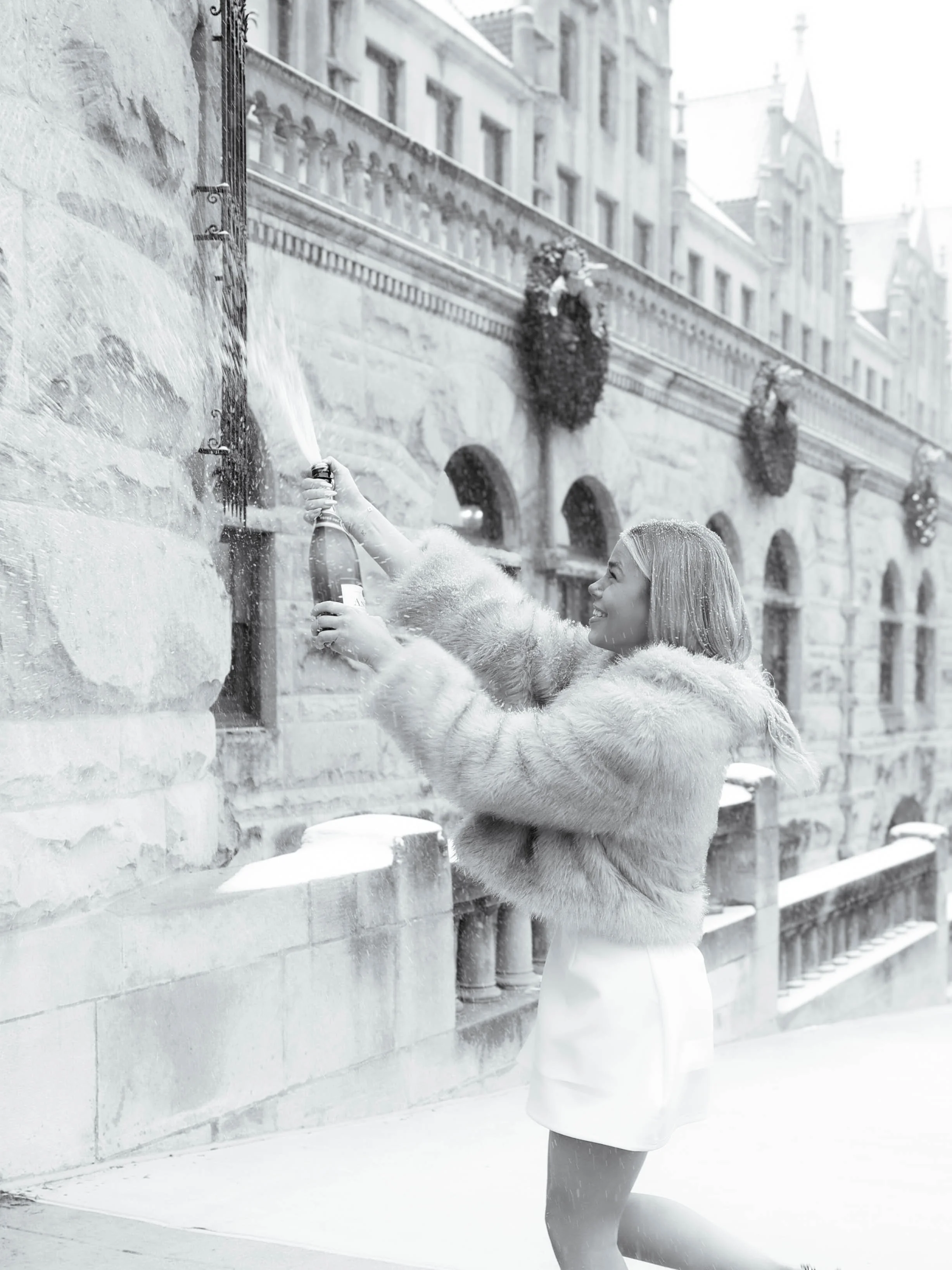 A woman in a fur coat holding a champagne bottle spraying it in celebration is in front of a historic stone building decorated with wreaths, during winter with snow.
