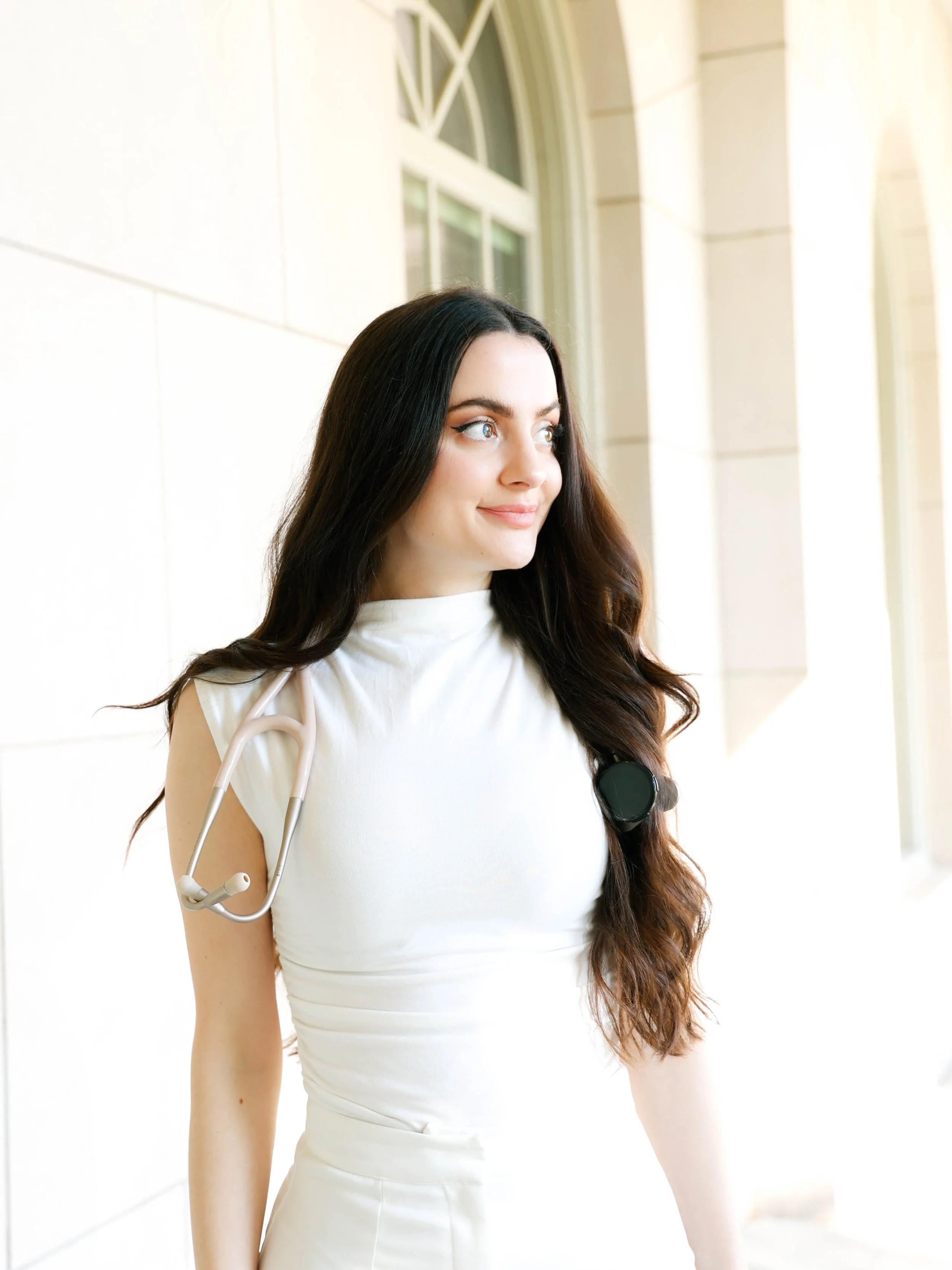 A young woman with long dark hair and blue eyes looking out a window, wearing a white sleeveless top and holding a stethoscope.