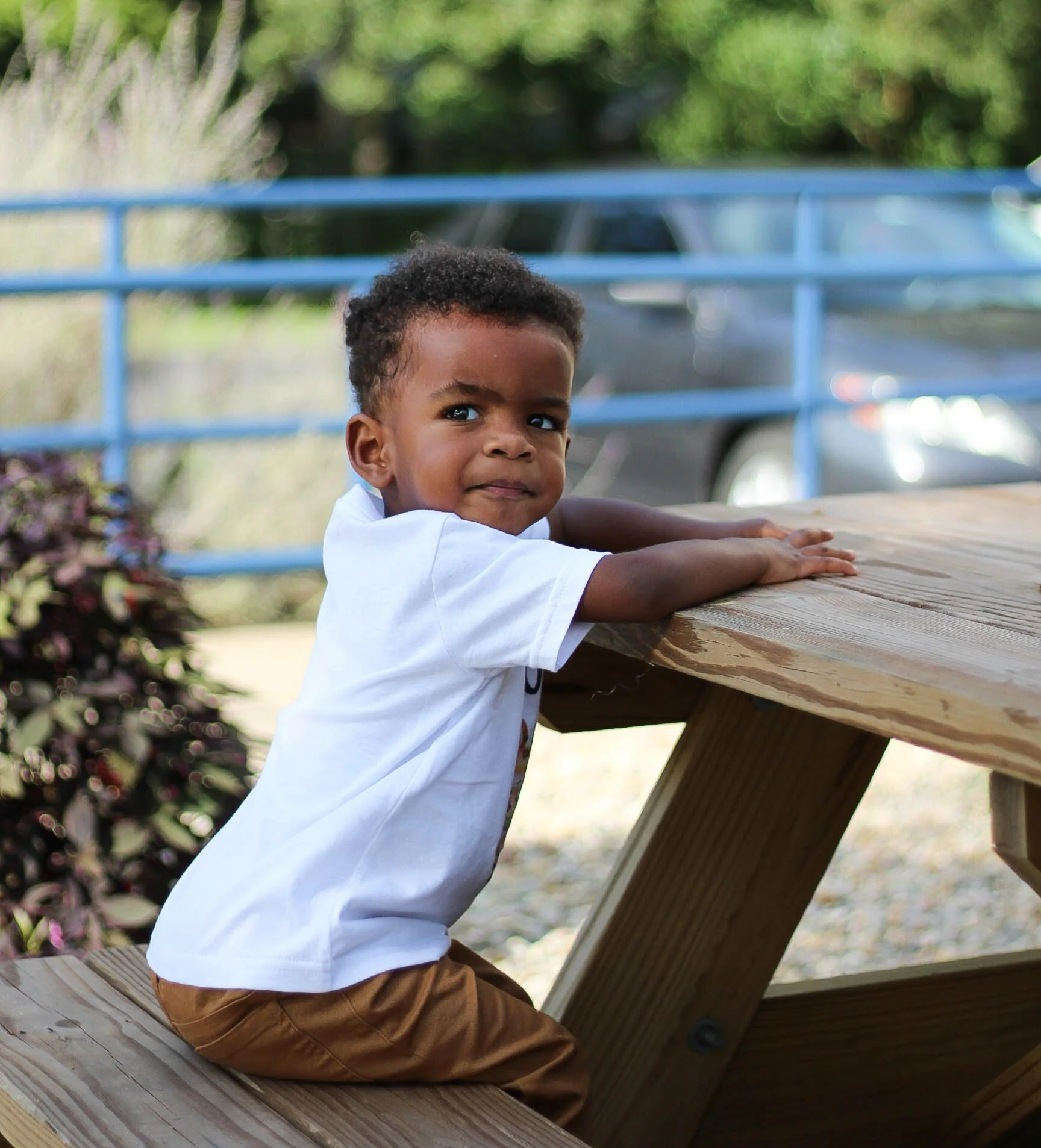 Young boy sitting at a wooden picnic table outdoors, leaning on his arms, looking towards the camera with a slight smile, with greenery and a parked car in the background.