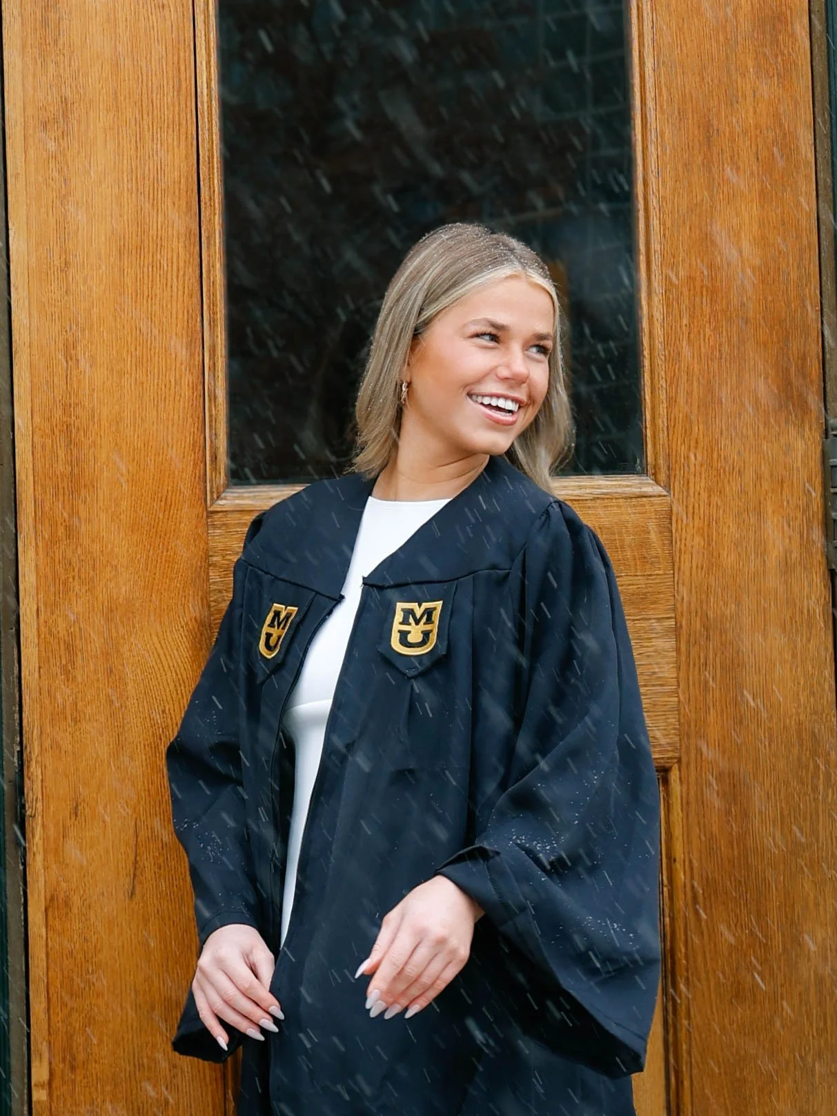 A young woman with shoulder-length blonde hair wearing a graduation gown with university patches, standing in front of a wooden door, smiling and looking to her right.
