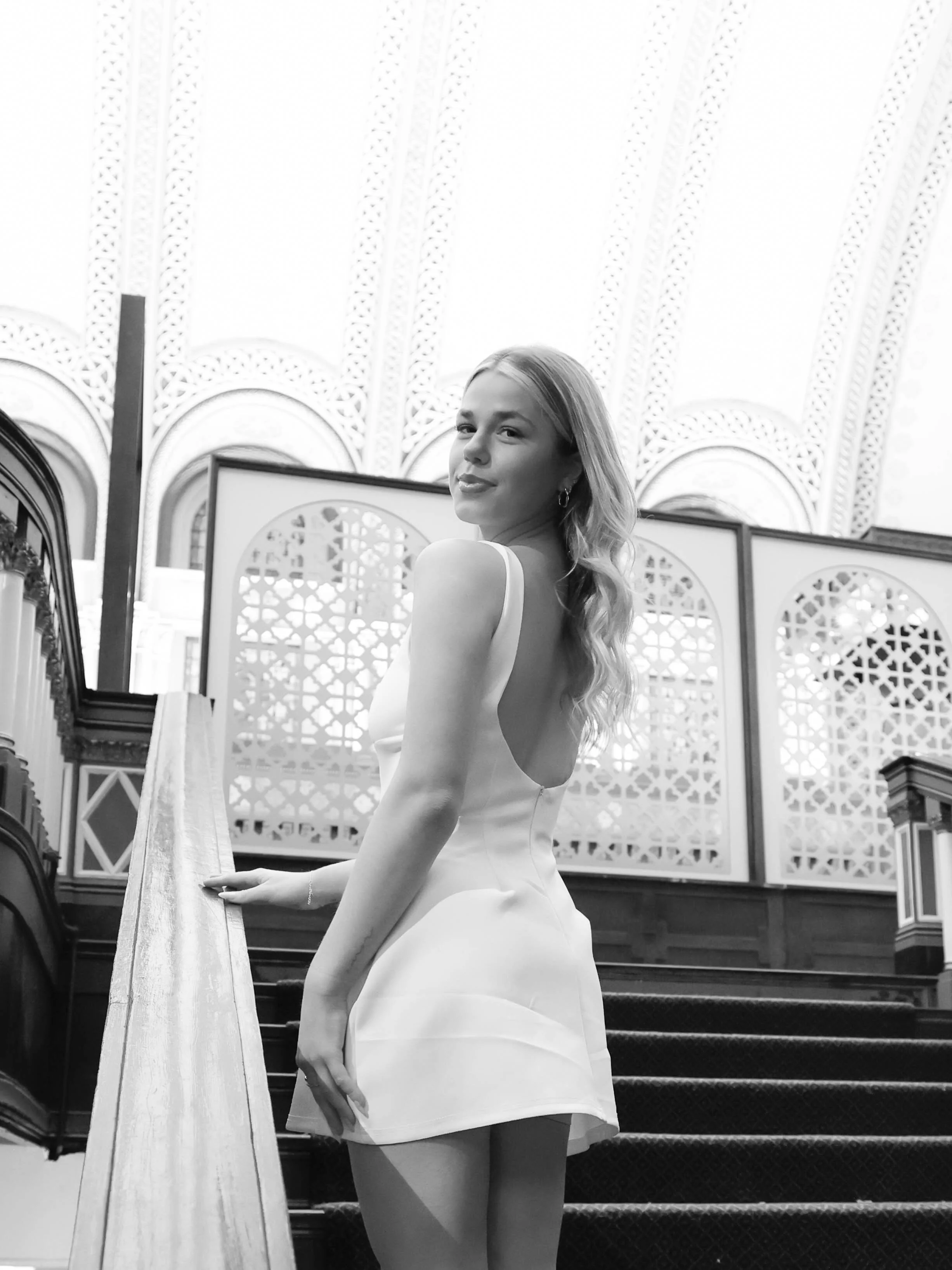 A woman with long wavy hair in a sleeveless white dress standing on a staircase in front of decorative folding screens in a bright, ornate room.
