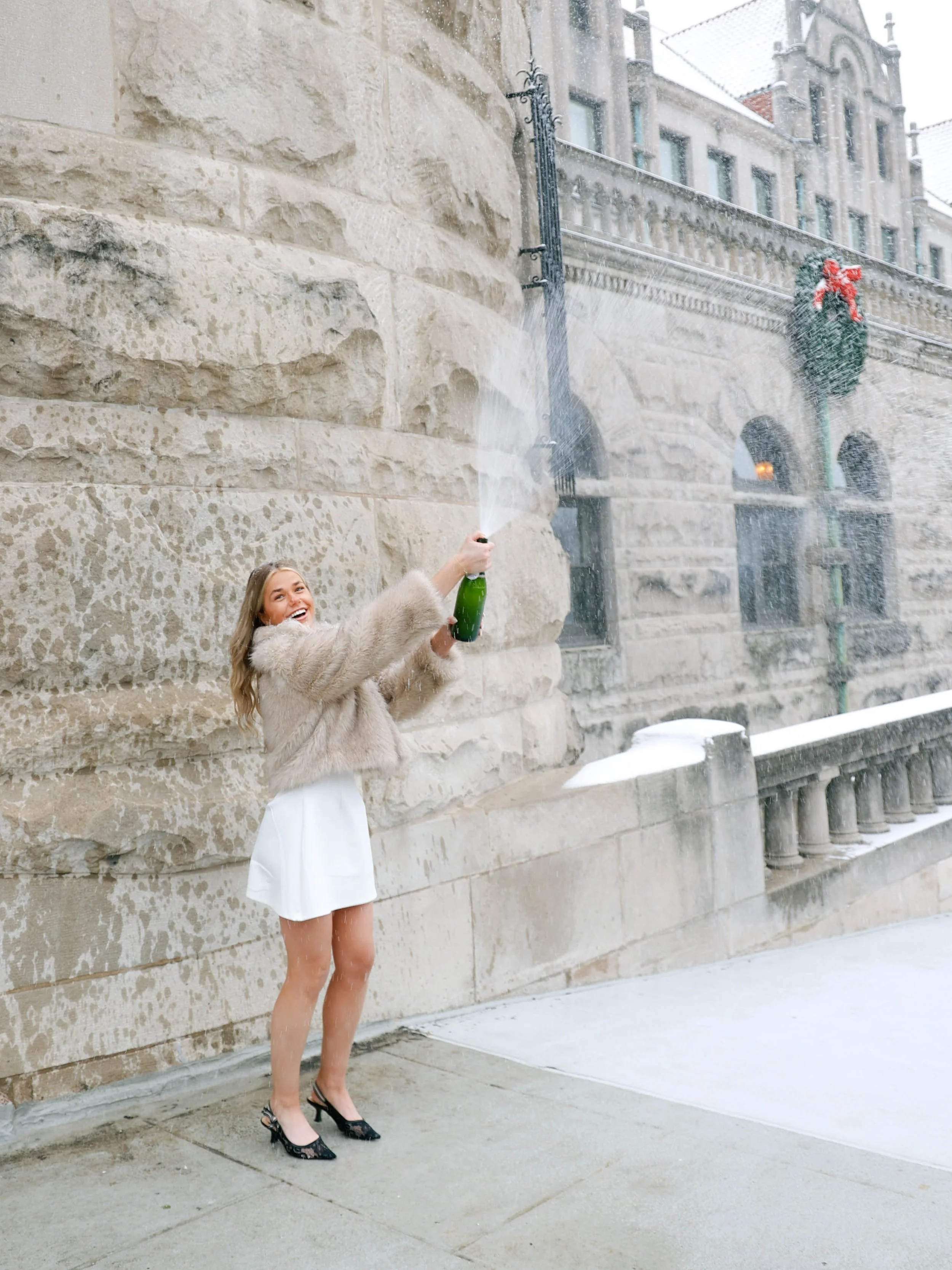 A woman standing outdoors on a wet sidewalk during winter, holding and opening a green champagne bottle with spray erupting out. She is wearing a beige fur coat, white skirt, and black high heels, with snow on the ground and a festive wreath hanging 