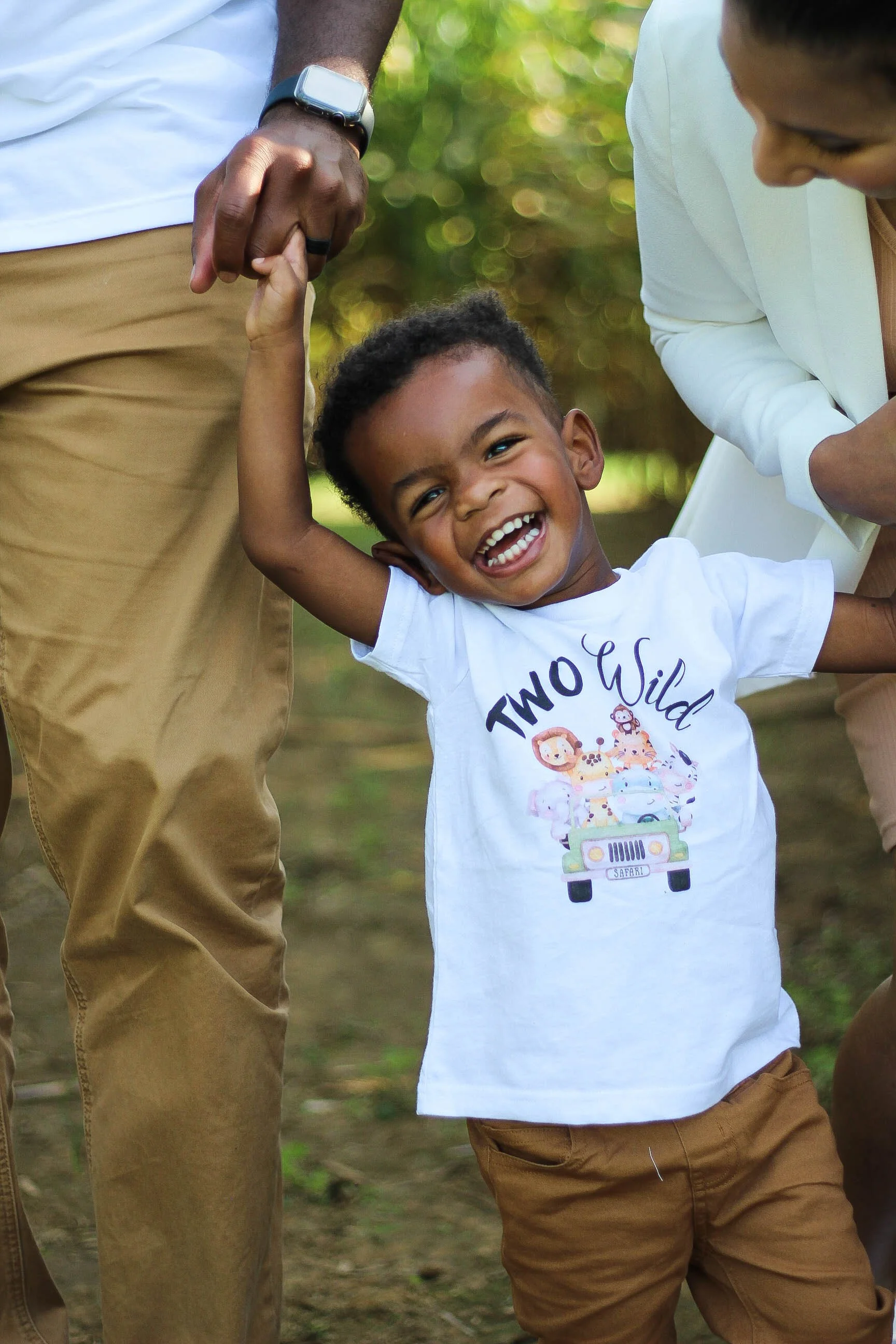 A young boy smiling and laughing, holding hands with an adult. The boy is wearing a white t-shirt with a cartoon safari vehicle and animal characters, and tan pants. The adult, partly visible, is wearing tan pants, a white shirt, and a smartwatch. Th
