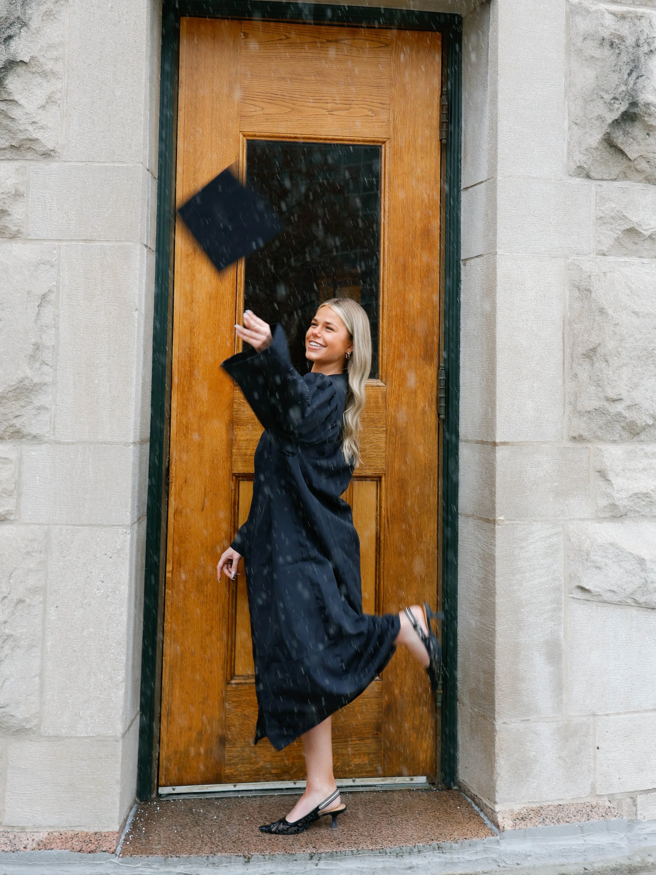 A woman in a black dress and high heels is standing and dancing on a doorstep in the rain, smiling and throwing a black purse into the air.