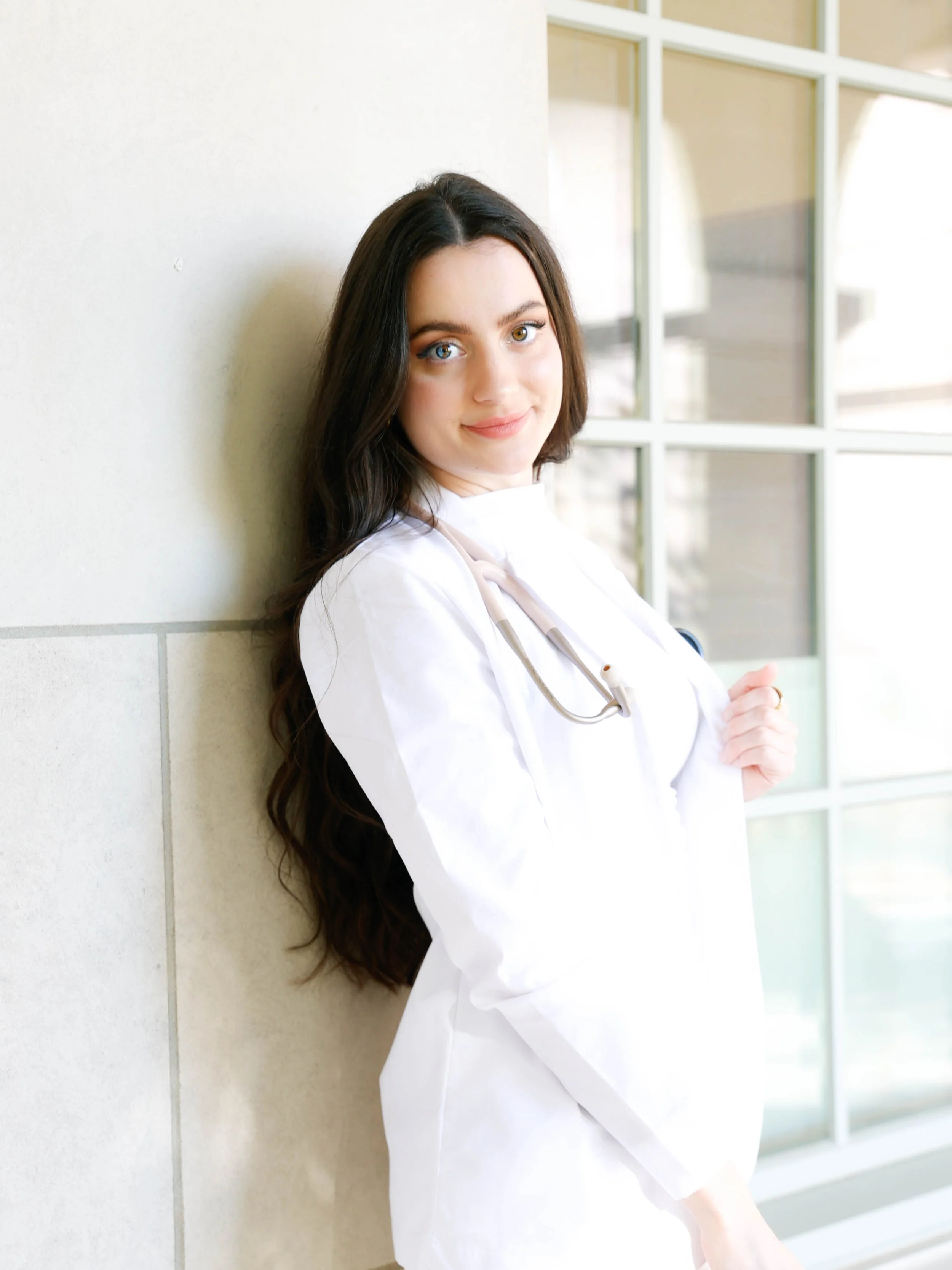 A young woman with long brown hair, blue eyes, and light skin leaning against a wall inside a building with large windows, wearing a white medical coat and a stethoscope around her neck.