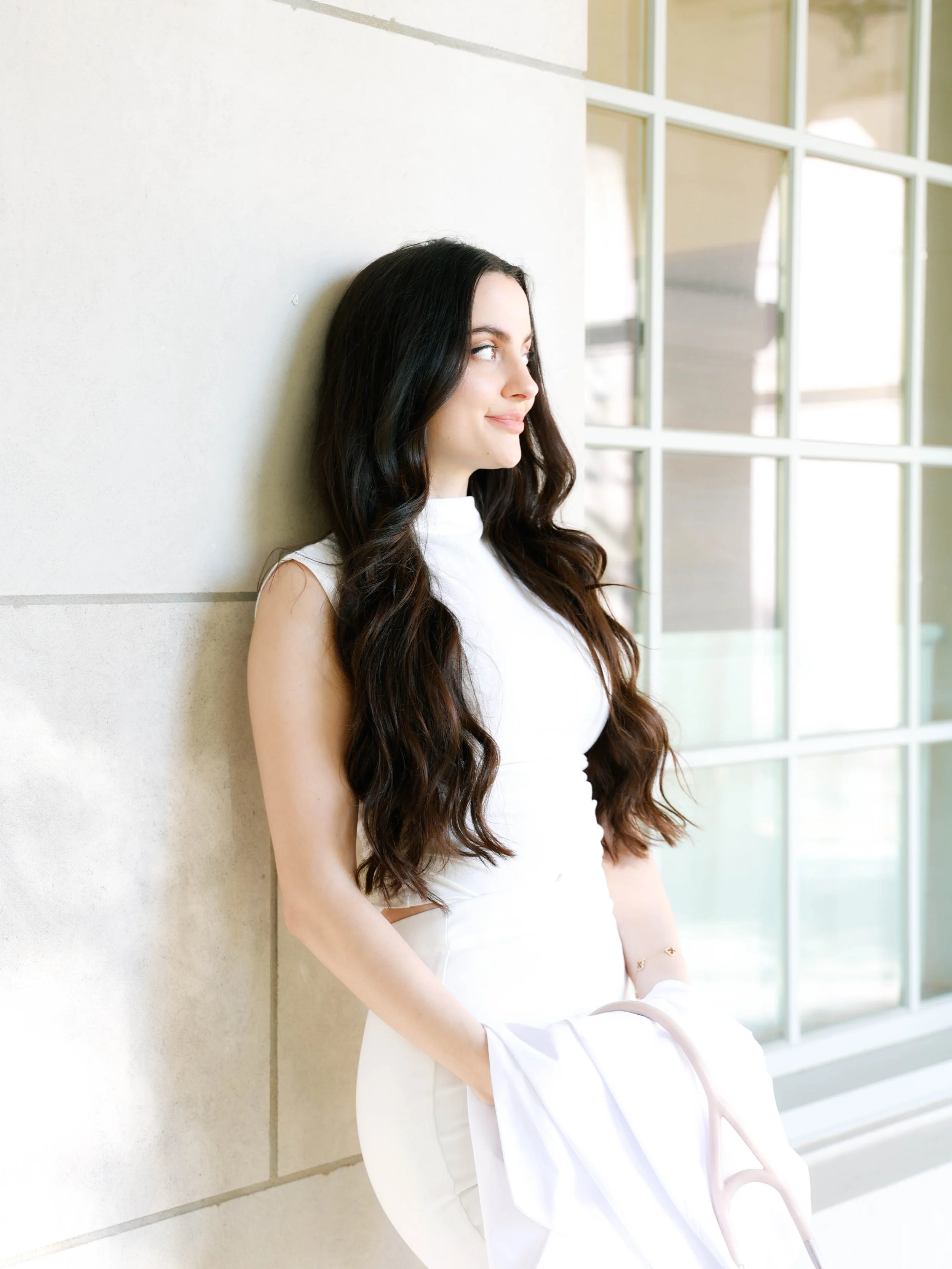 Young woman with long dark wavy hair leaning against a wall near large windows, smiling and looking to the side, dressed in a white sleeveless top and holding a light pink bag.