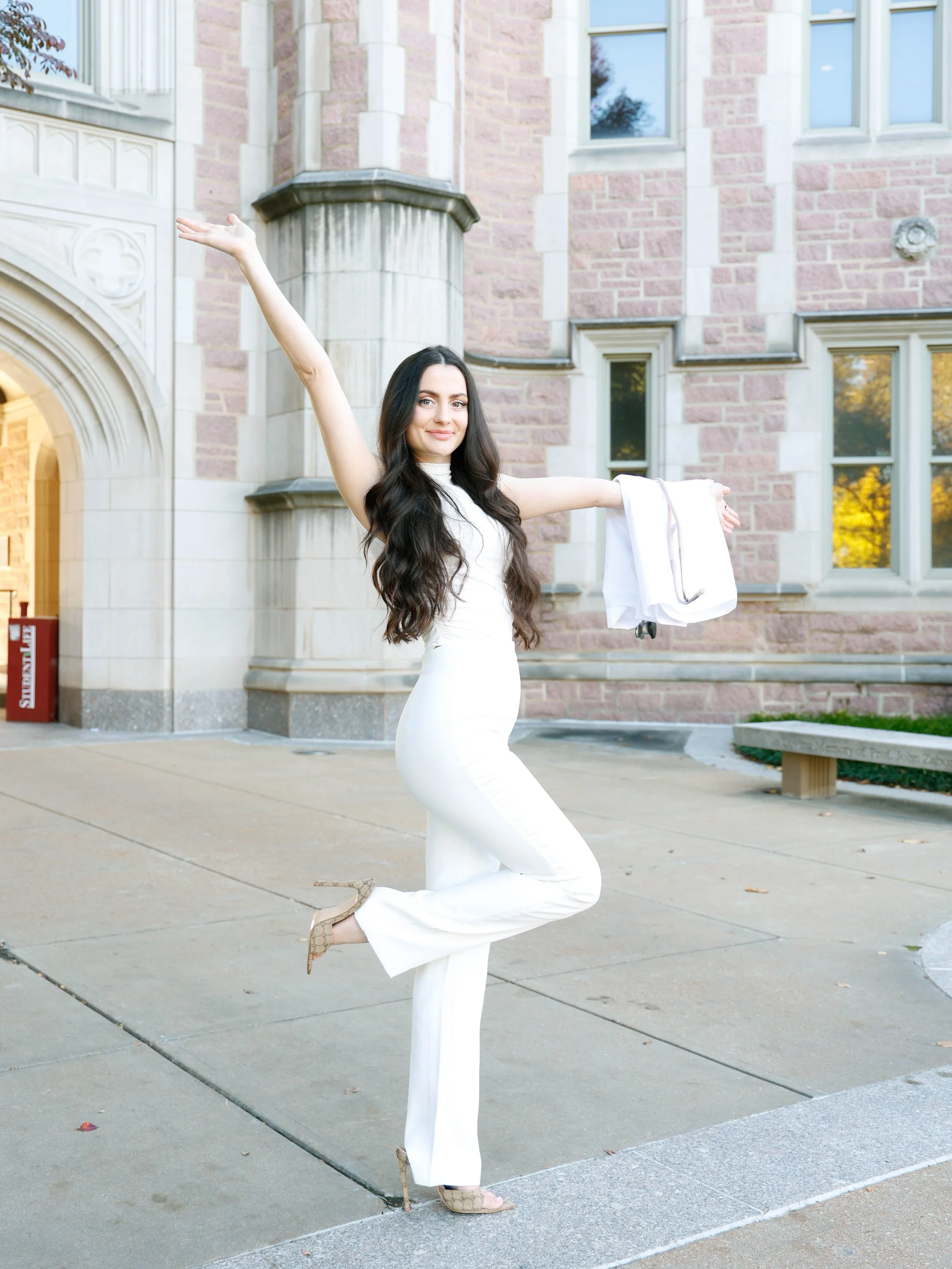 A woman in a white dress and high heels is balancing on one leg with her arms raised, outside a brick building with large windows and an arched entrance.