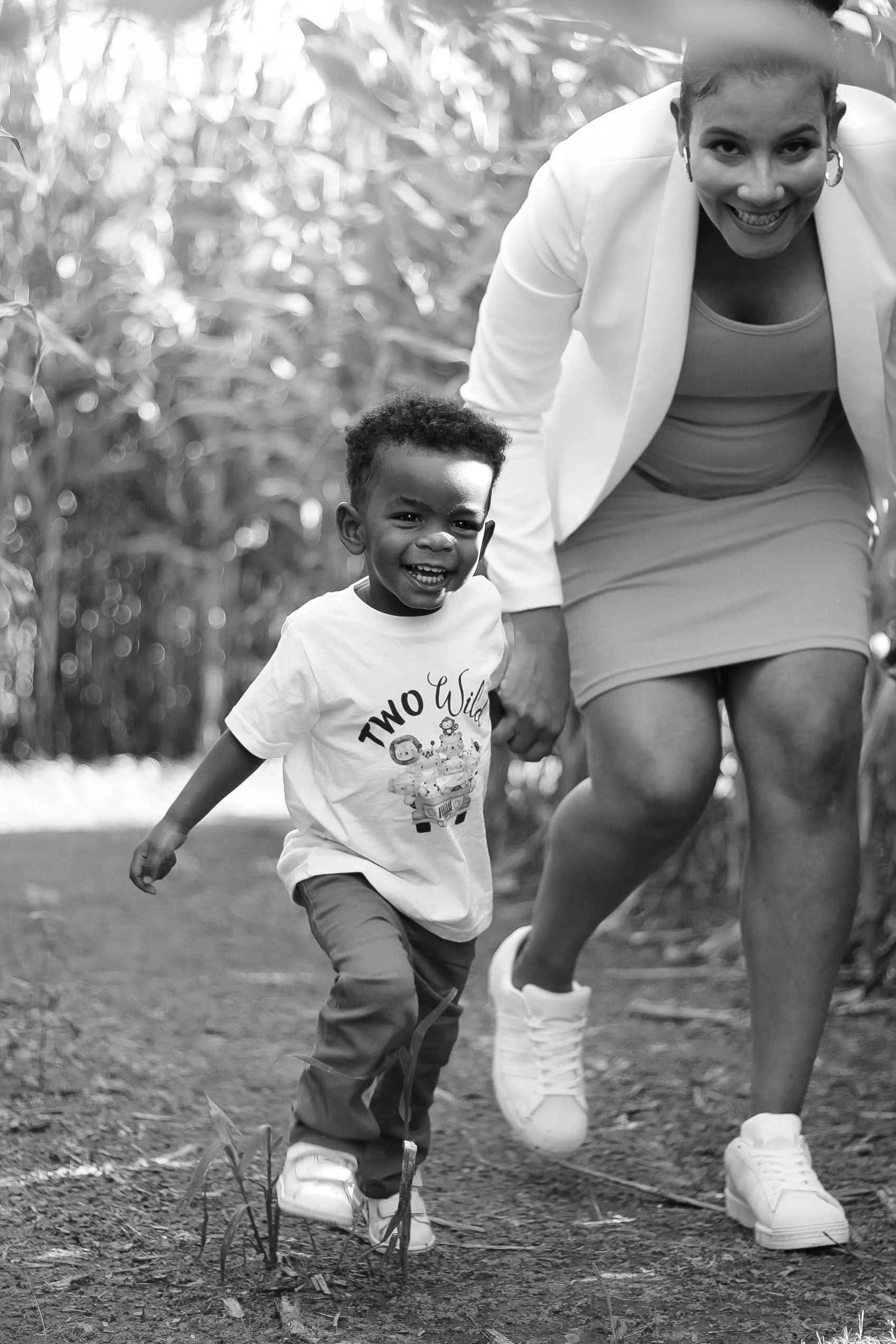 A woman and a young boy running outdoors on a dirt path with tall grass or plants in the background. The woman is smiling and holding the boy's hand, and both appear joyful.