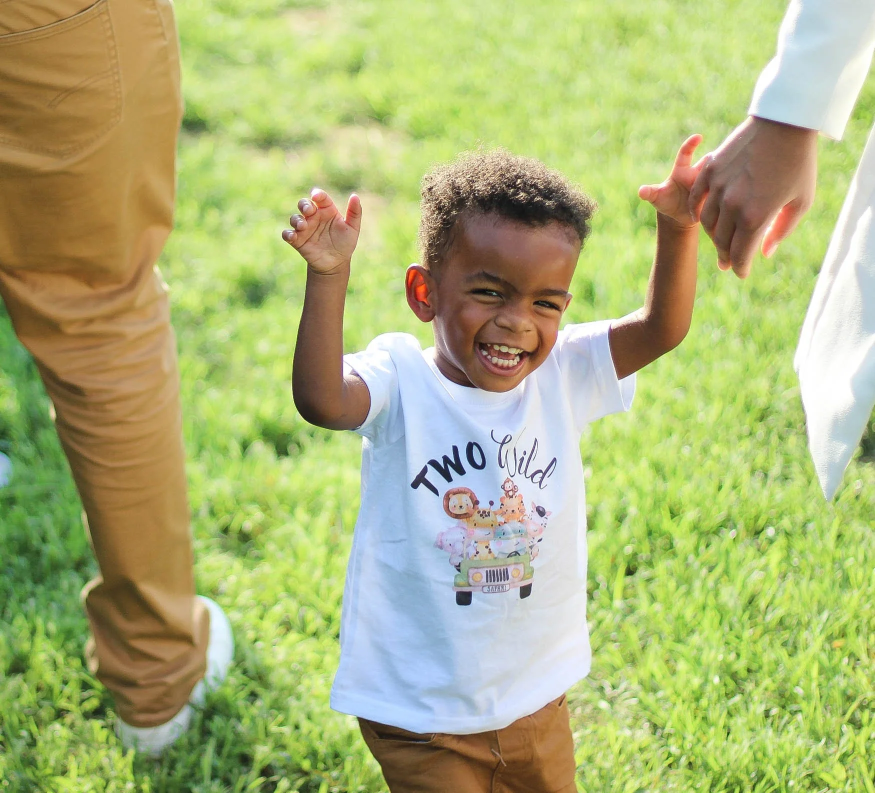 A happy young boy with curly hair smiling and raising his hands while holding hands with an adult outdoors on a grassy field.