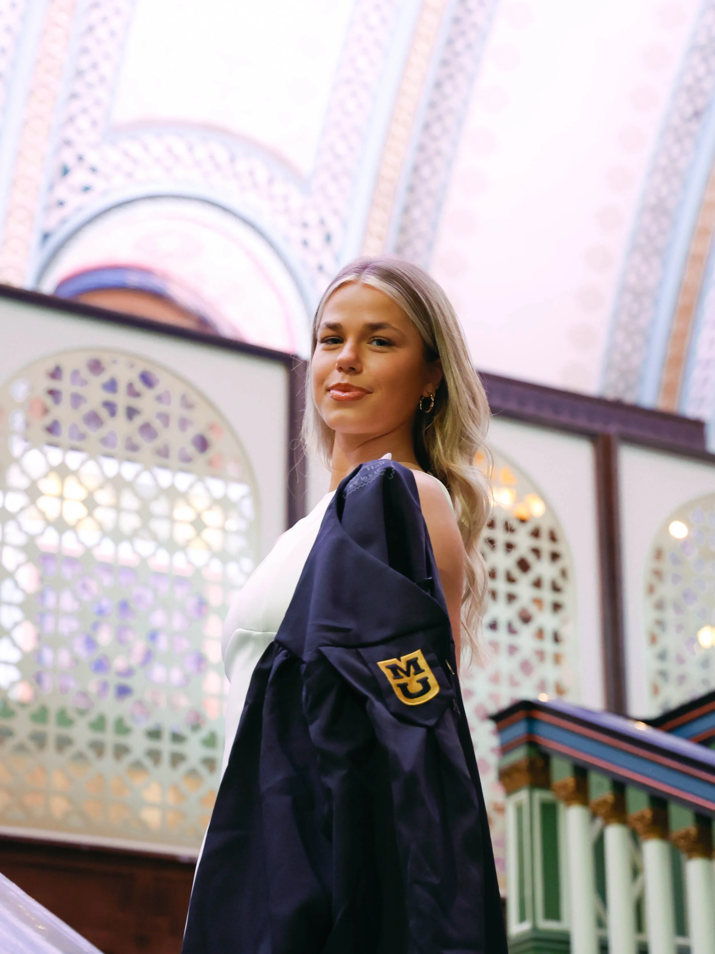 A young woman with blonde hair wearing a graduation gown with a Michigan emblem, smiling inside a building with ornate ceiling decorations and vibrant architectural details.