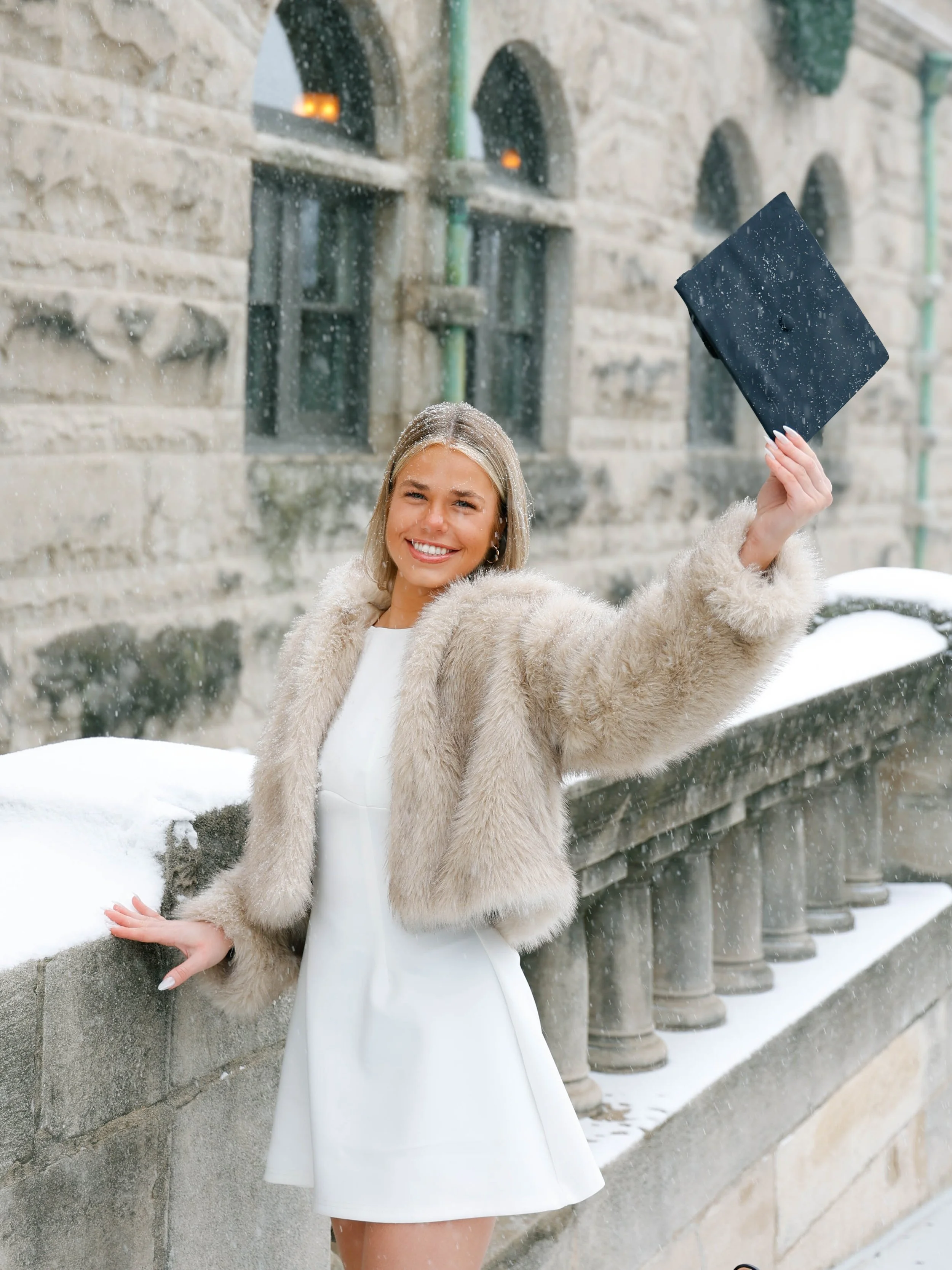 A smiling woman in a fur coat and white dress holding a snowball while standing outdoors near a stone wall with arched windows in snowy weather.