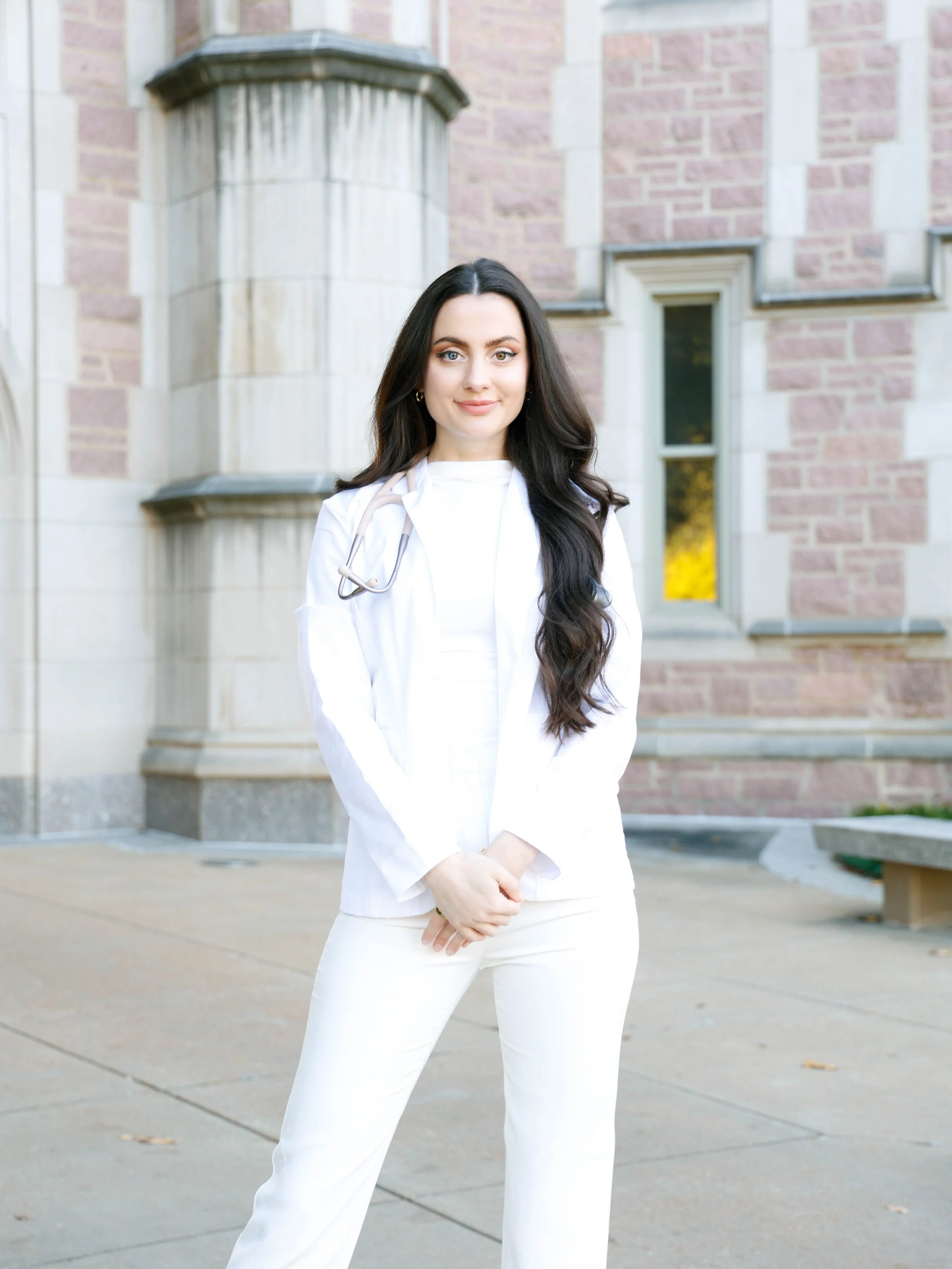 A young woman in a white medical coat stands outdoors in front of a brick building, with a stethoscope around her neck and her hands folded in front of her.