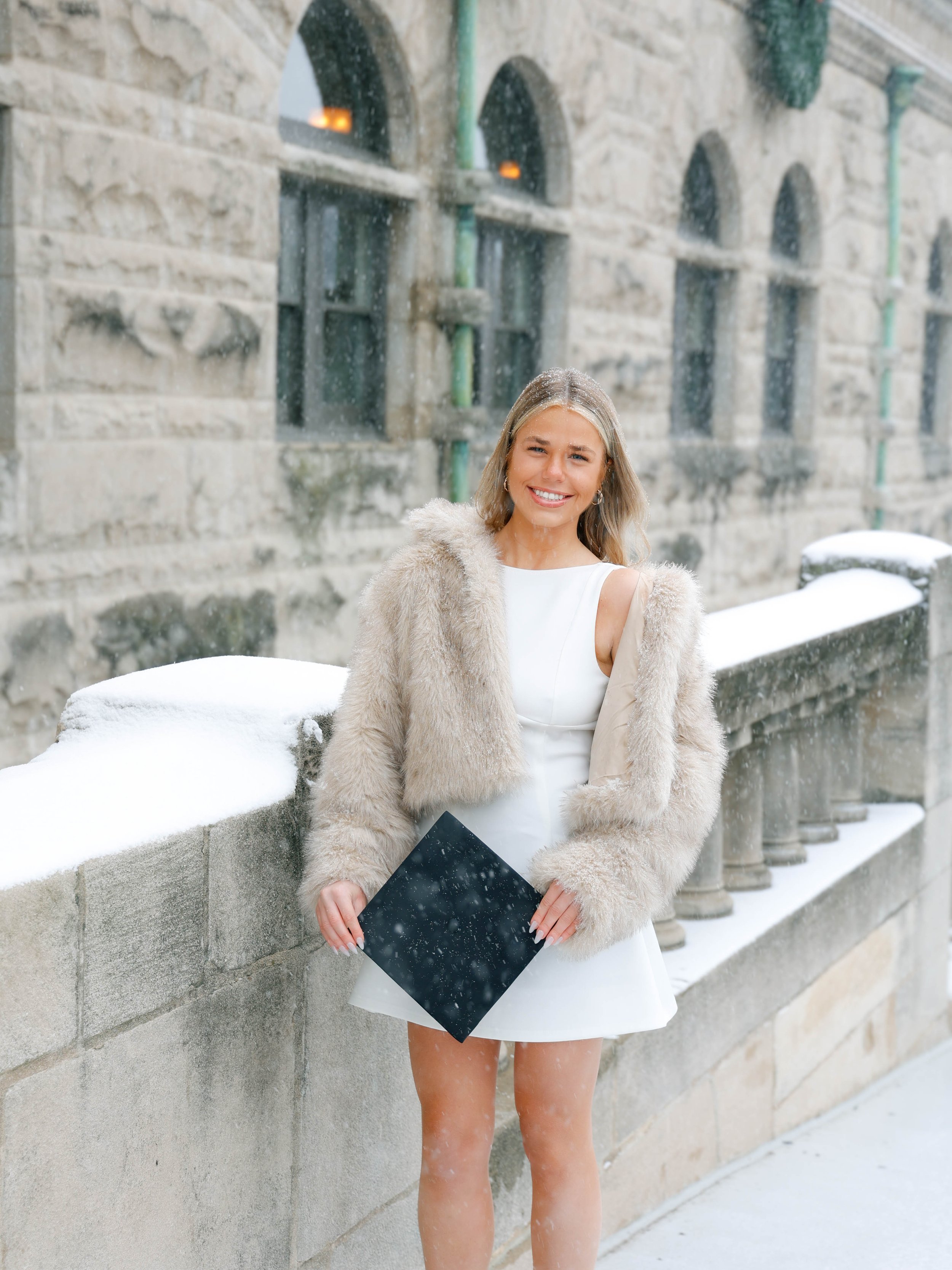Woman smiling outside in cold weather, wearing a white dress and a beige fur coat, holding a black folder, with snow on the ground and on a stone wall behind her.
