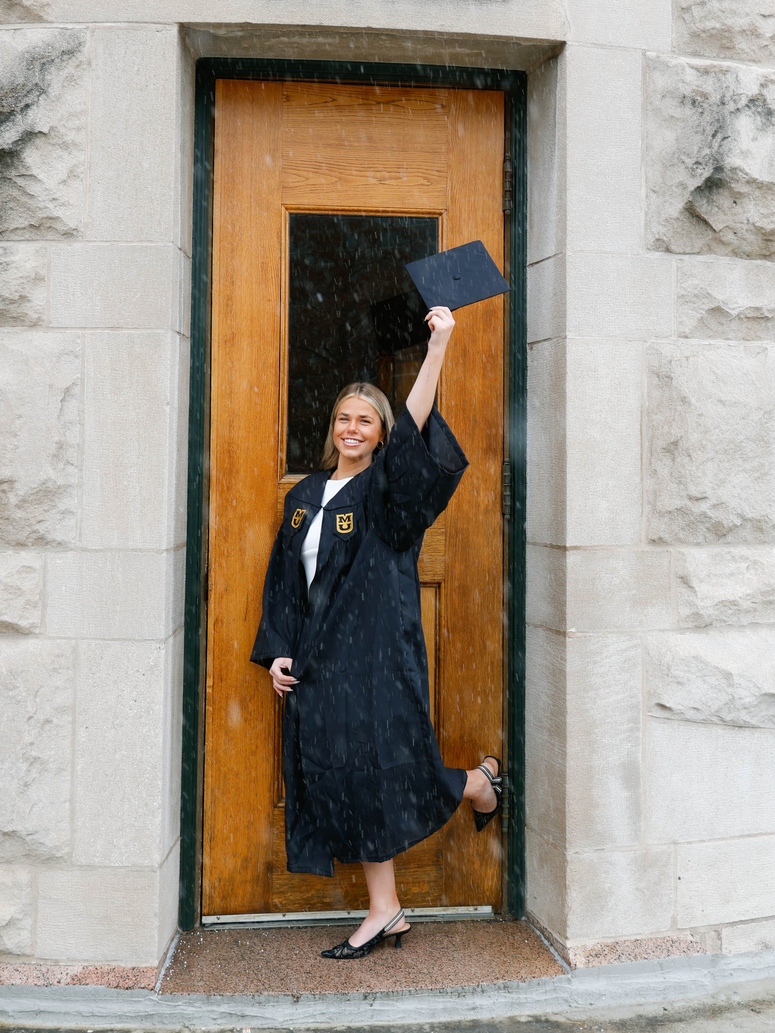 A smiling young woman in a graduation gown holding her diploma above her head while standing in a doorway during rain.