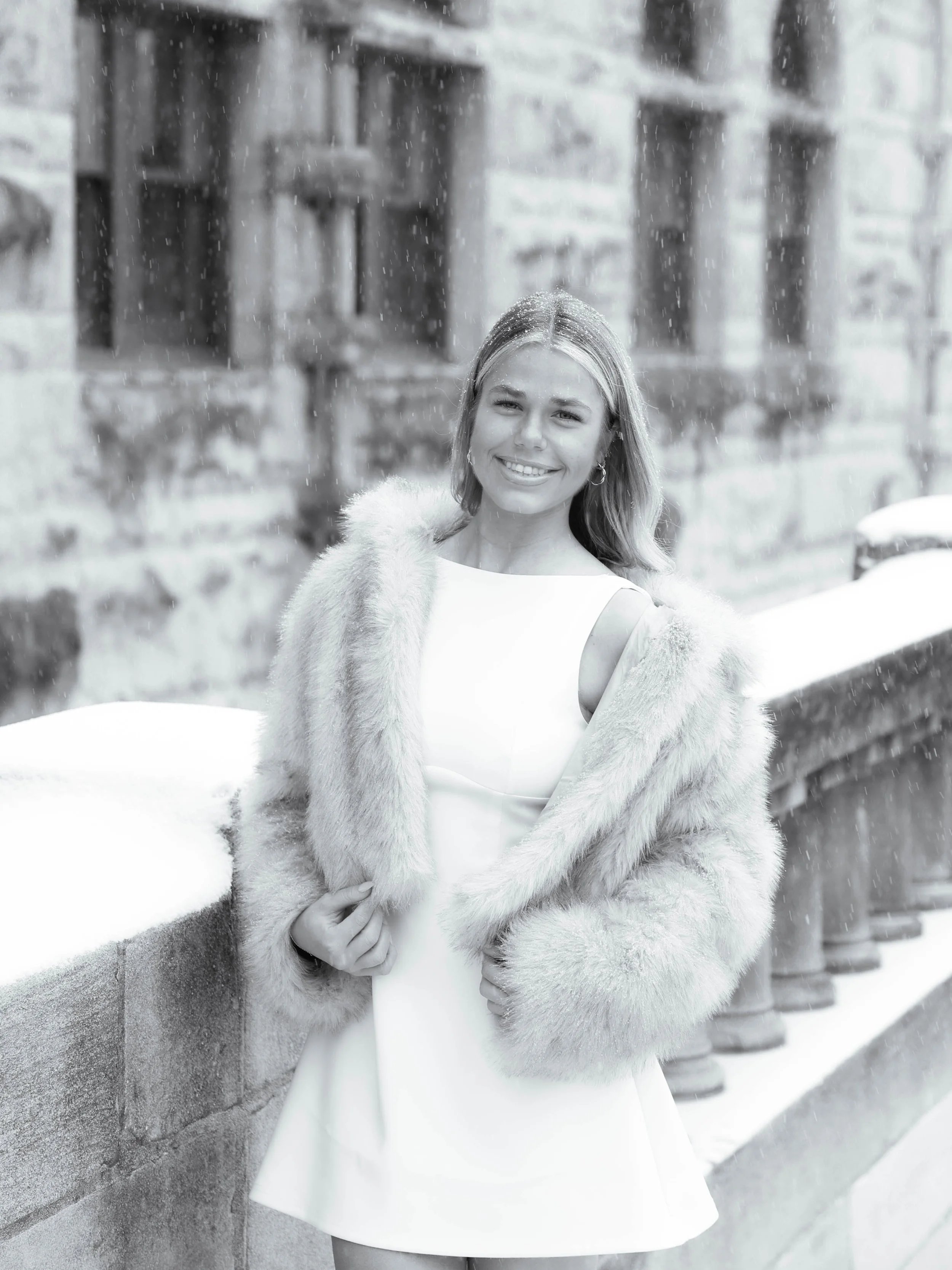 A smiling woman wearing a white dress and a fur coat standing outdoors in a snowy environment, leaning against a concrete wall on a snowy street with buildings and a car in the background.