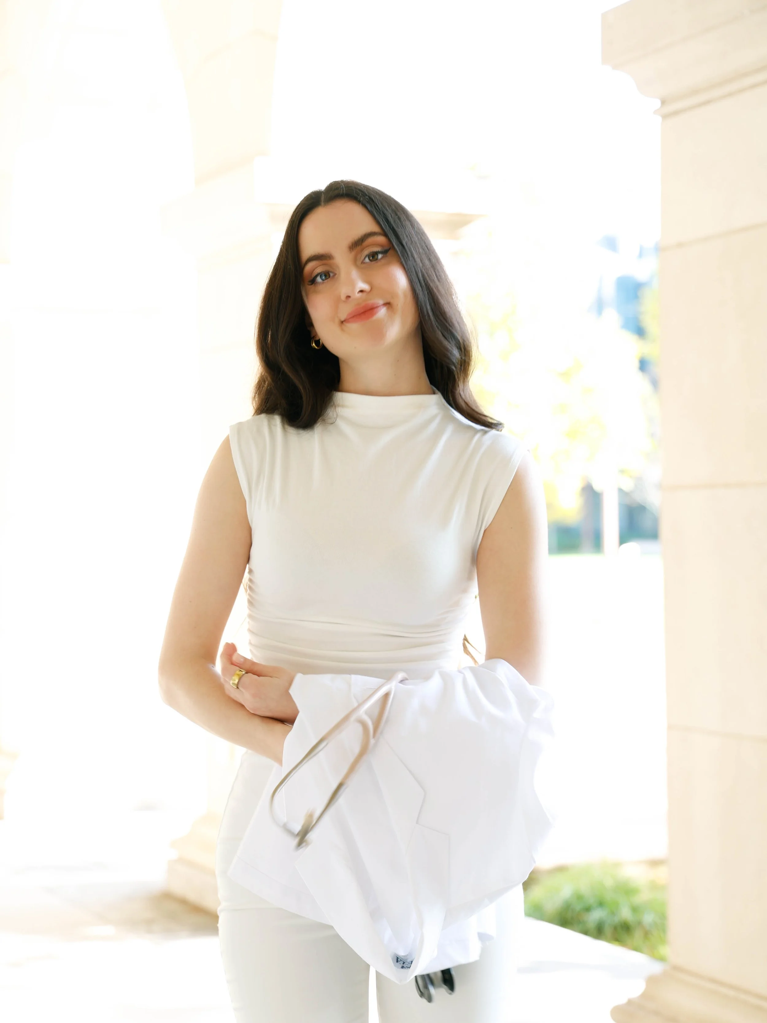 A woman with dark hair, blue eyes, and light skin standing outdoors near a light-colored building, wearing a sleeveless white top, and smiling while holding a white jacket.