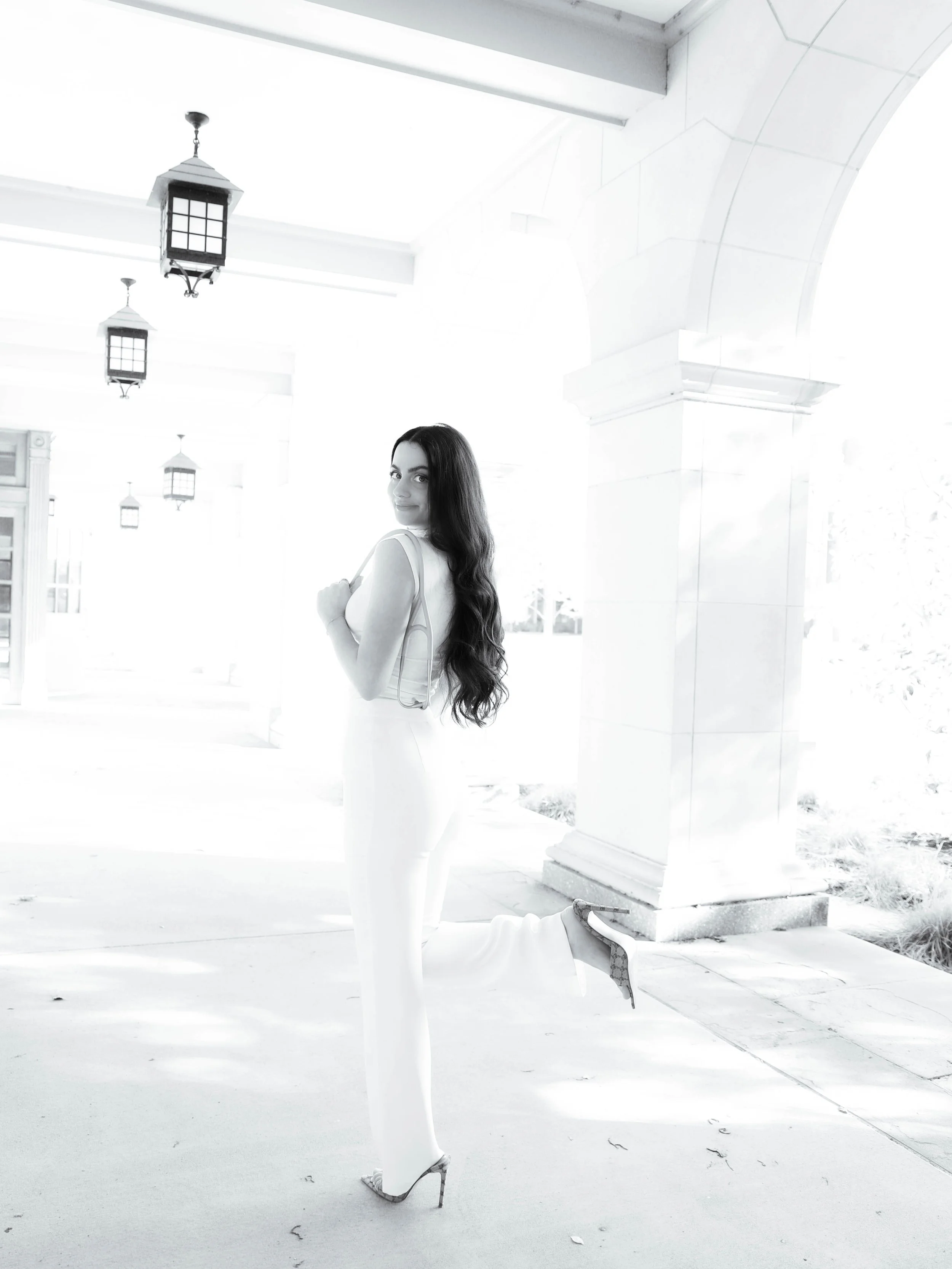 A woman with long wavy hair and high heels kneeling outdoors beside a white column under a porch, with trees and a bright sky in the background.