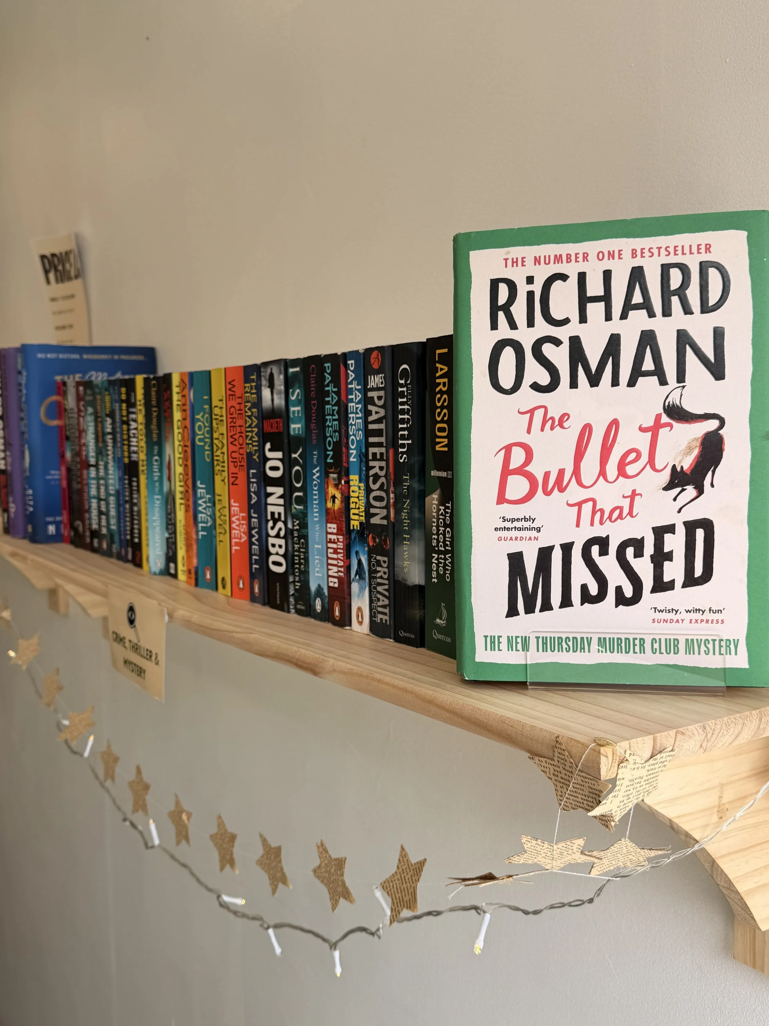 A wooden shelf holding a collection of books with a prominent book titled 'The Bullet That Missed' by Richard Osman. The shelf is decorated with a string of star-shaped fairy lights.