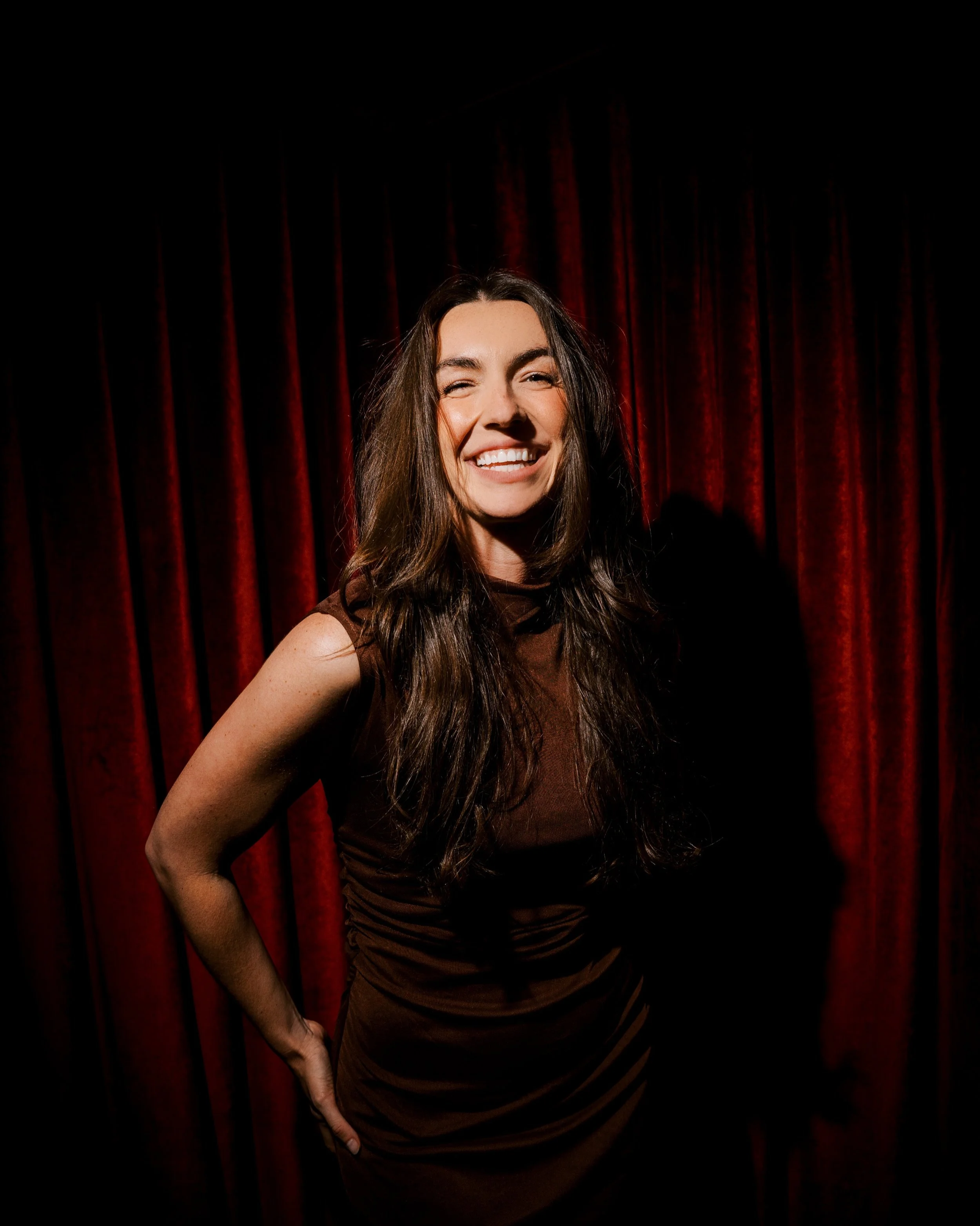 A young woman with long brown hair, smiling brightly, standing in front of a red velvet curtain. She is wearing a sleeveless, dark brown dress and has her left arm bent with her hand on her hip.
