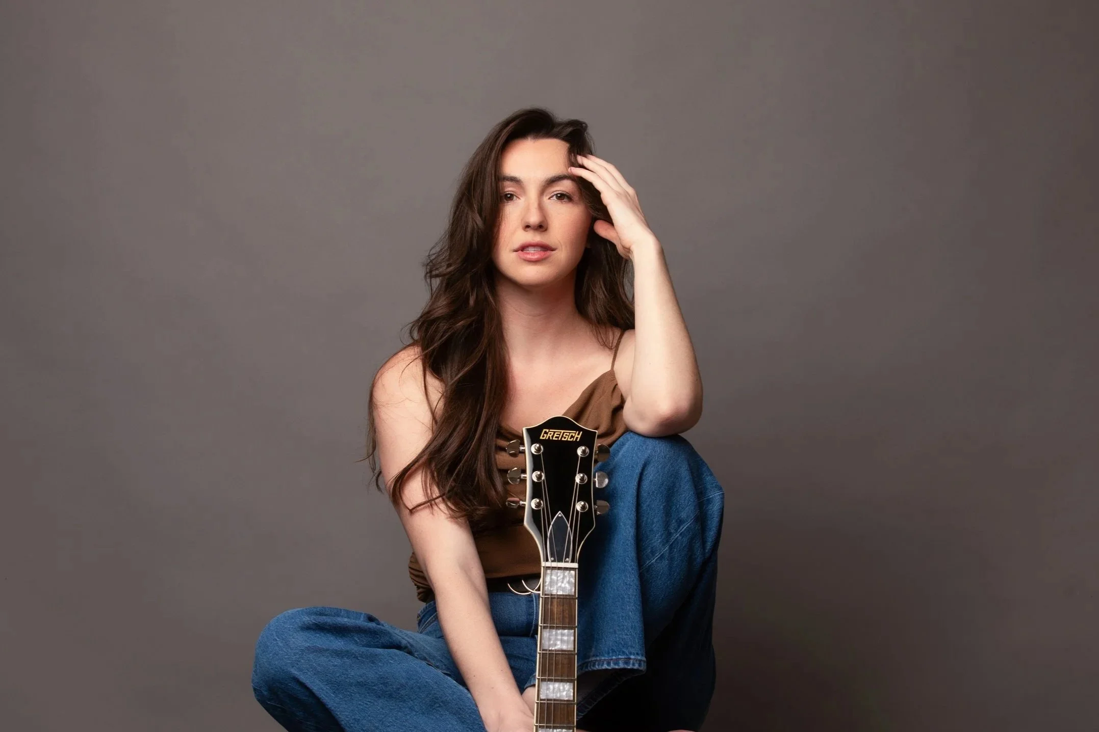 A woman with long wavy brown hair, wearing a brown top and blue jeans, sitting cross-legged against a plain gray background, holding an acoustic guitar with a 'GRETSCH' logo visible on the headstock, her left hand resting on her forehead in a relaxed
