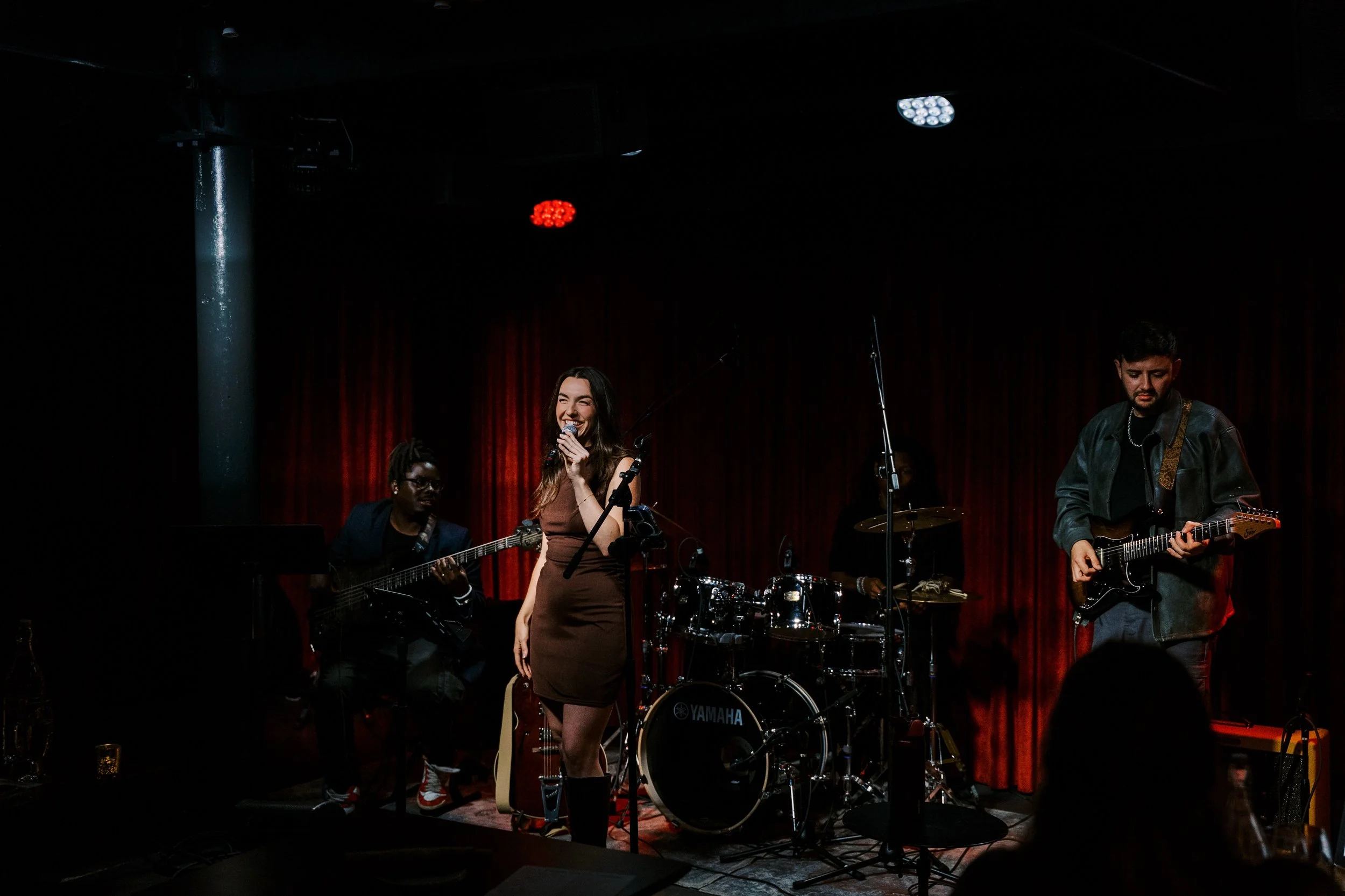 A live music performance on a stage with a female singer and several musicians, including guitarists and a drummer, under dim lighting with red curtains in the background.