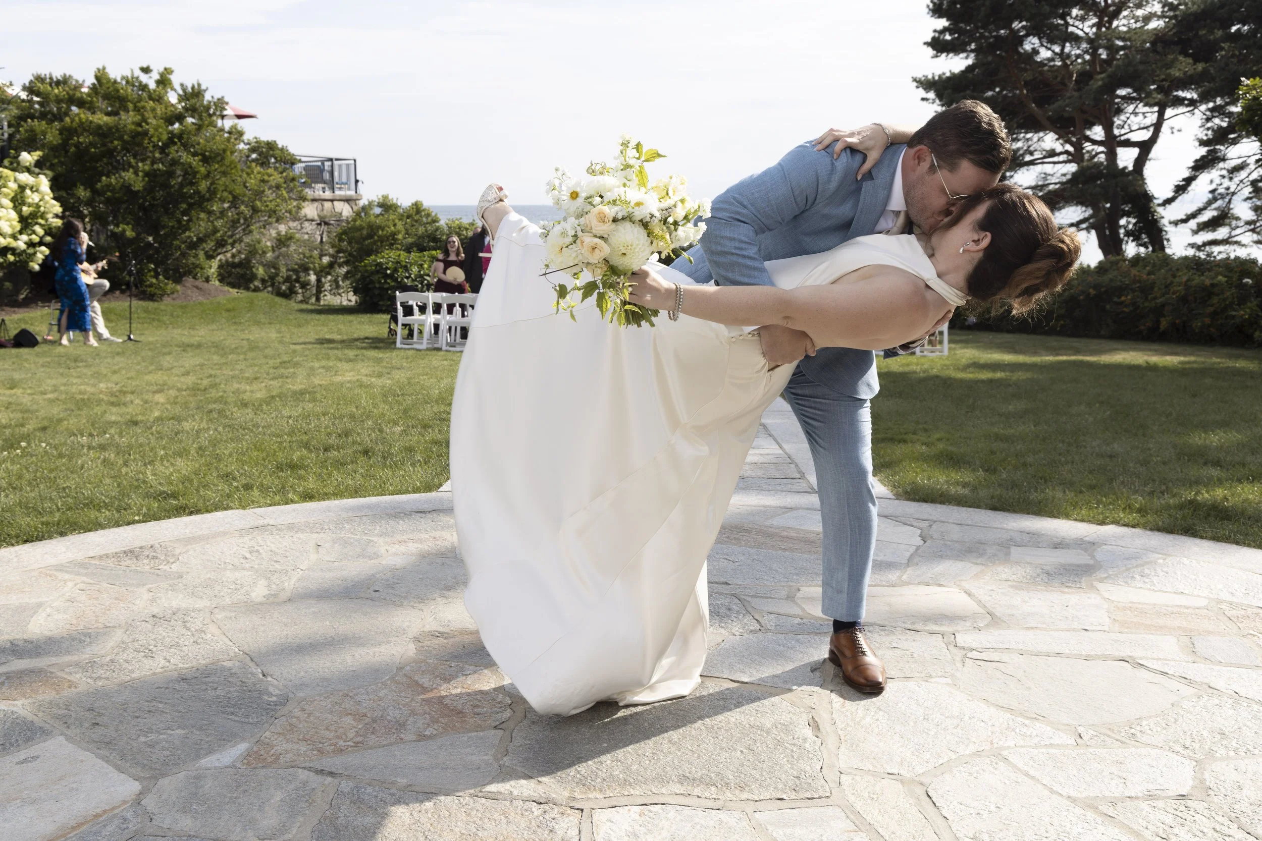 A couple dancing at an outdoor wedding ceremony, with a bouquet of flowers in the bride's hand, in a green garden with trees and a few guests in the background.