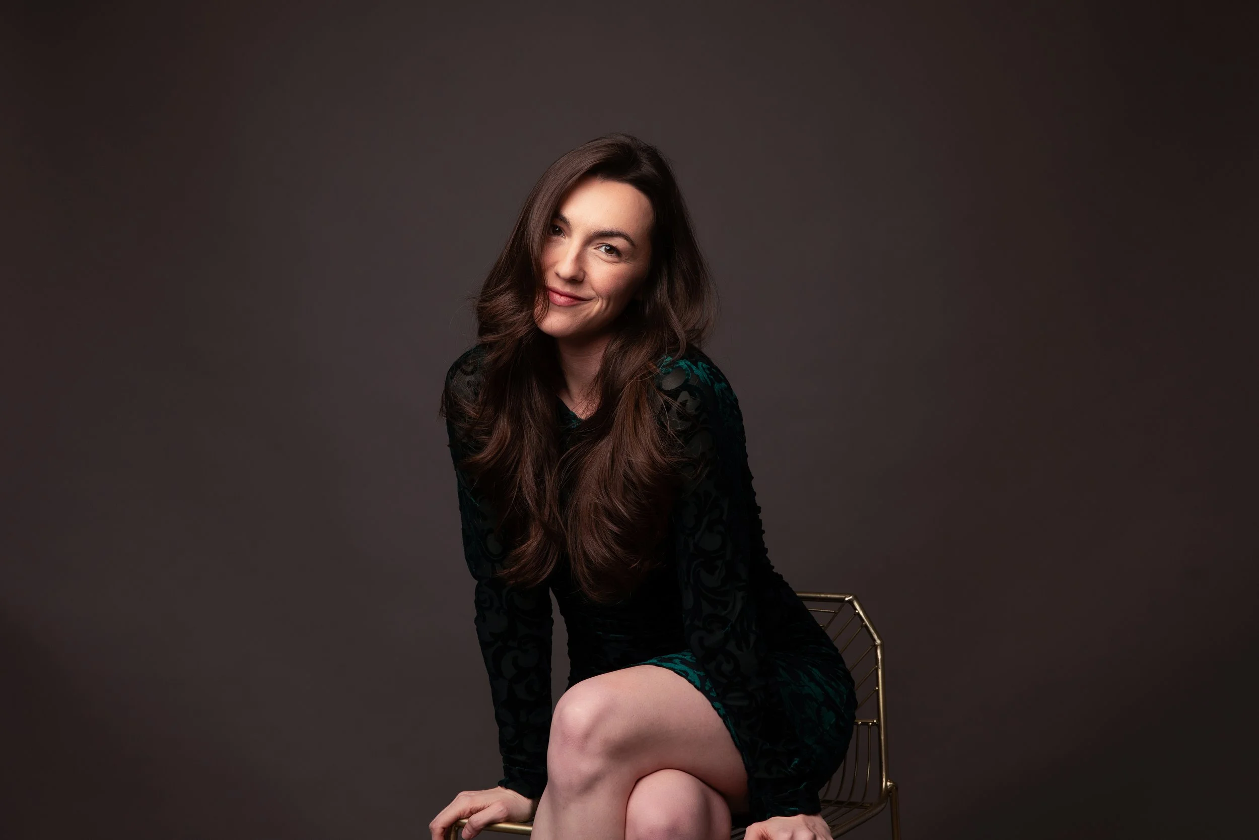 A woman with long wavy brown hair sitting on a gold wire chair, wearing a dark velvet dress with turquoise accents, against a dark gray background.