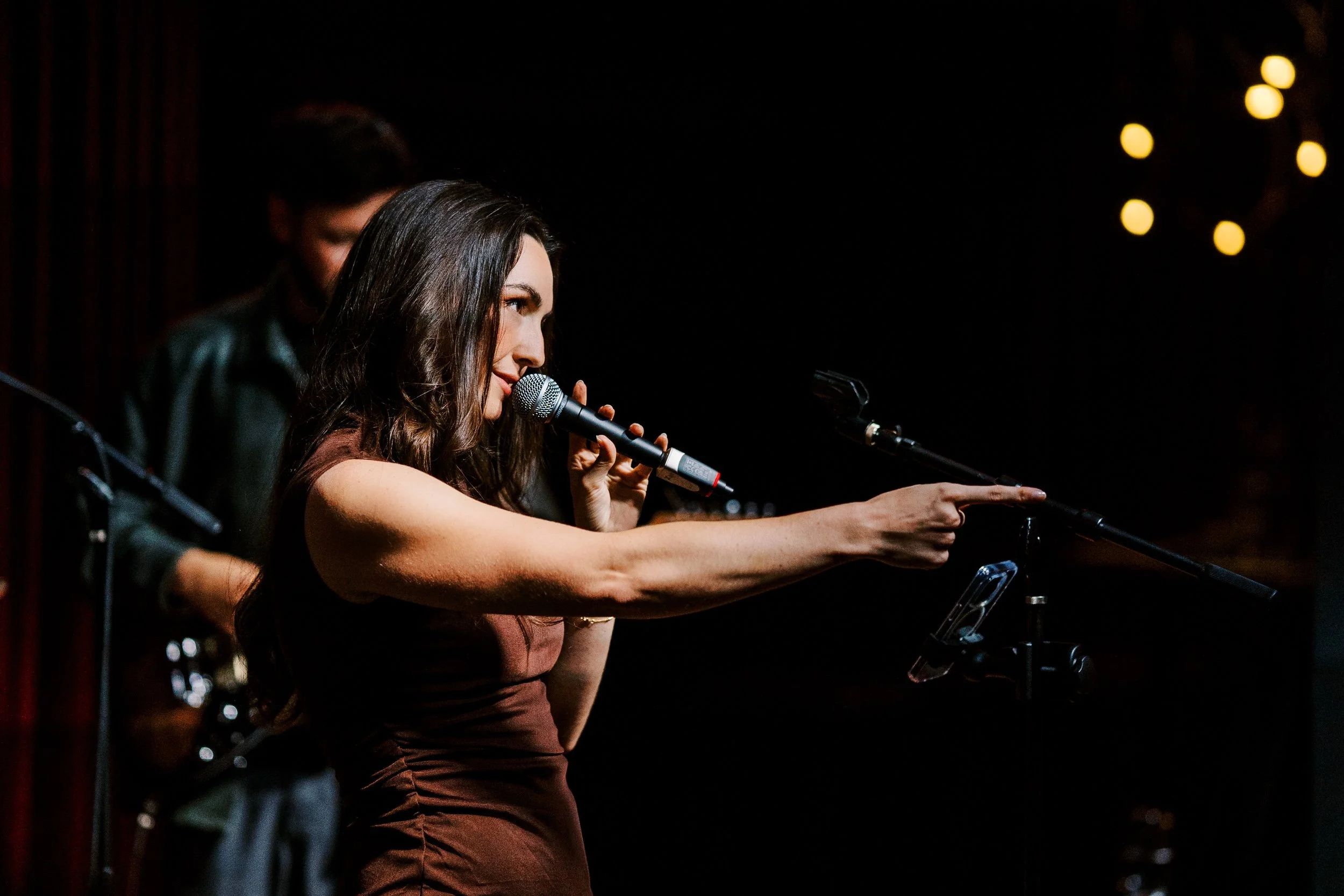 Woman singing or speaking into a microphone on stage, with a person in the background, against a dark background with round yellow stage lights.