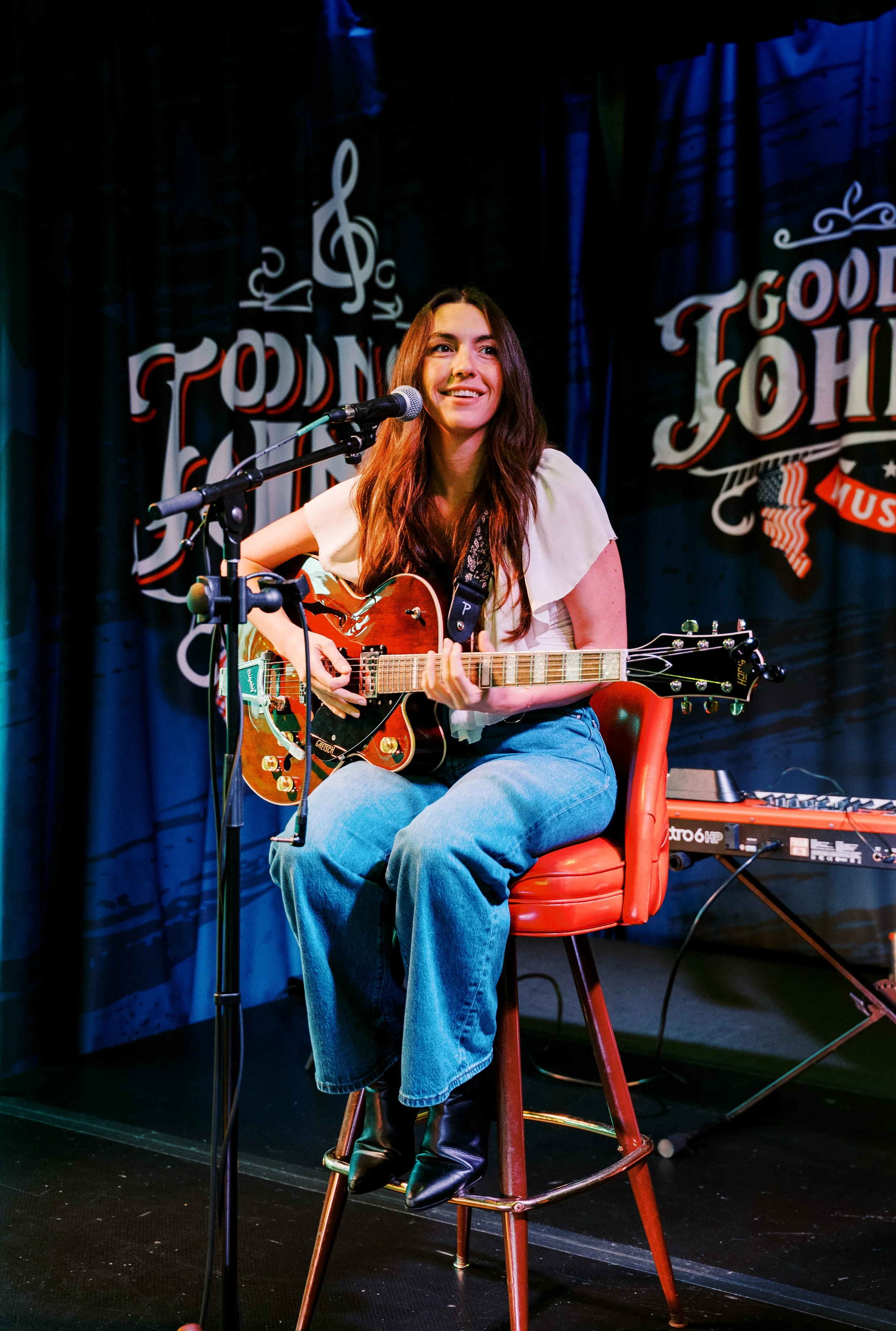 A young woman with long brown hair, wearing a white top and blue jeans, sits on a red stool on stage, playing an electric guitar and singing into a microphone. The background features a black curtain with music-themed writing and symbols.