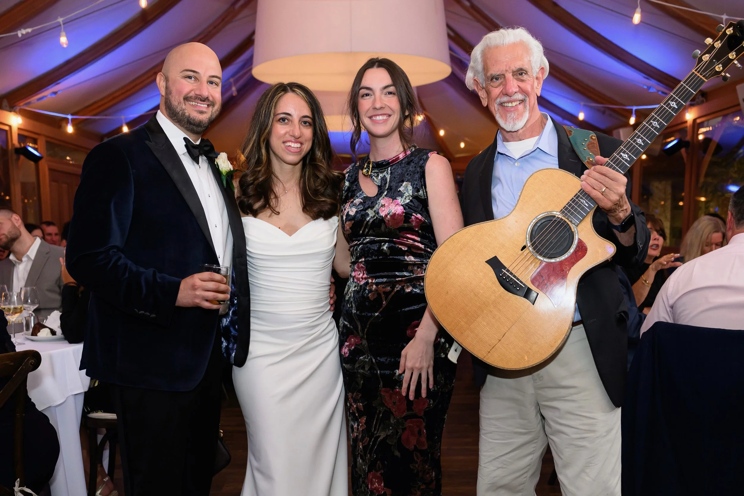Group of four people, three women and one man, standing close together at a social event with decorated interior, smiling at the camera. The man on the right is holding an acoustic guitar.