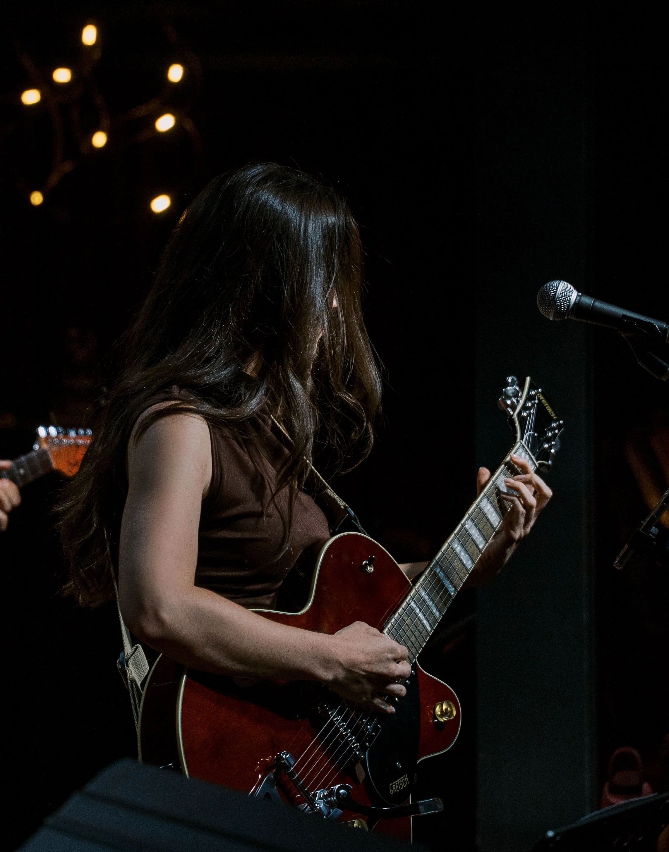 A woman with long brown hair playing an electric guitar on stage, with a microphone nearby and a dark background with yellow stage lights.