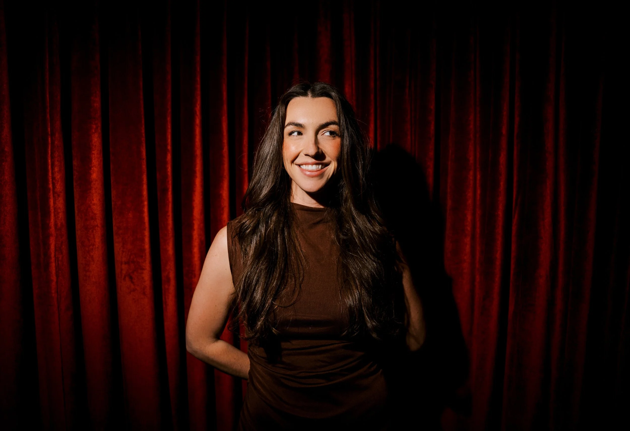 A woman with long dark hair smiling and looking to her right in front of red velvet curtains.