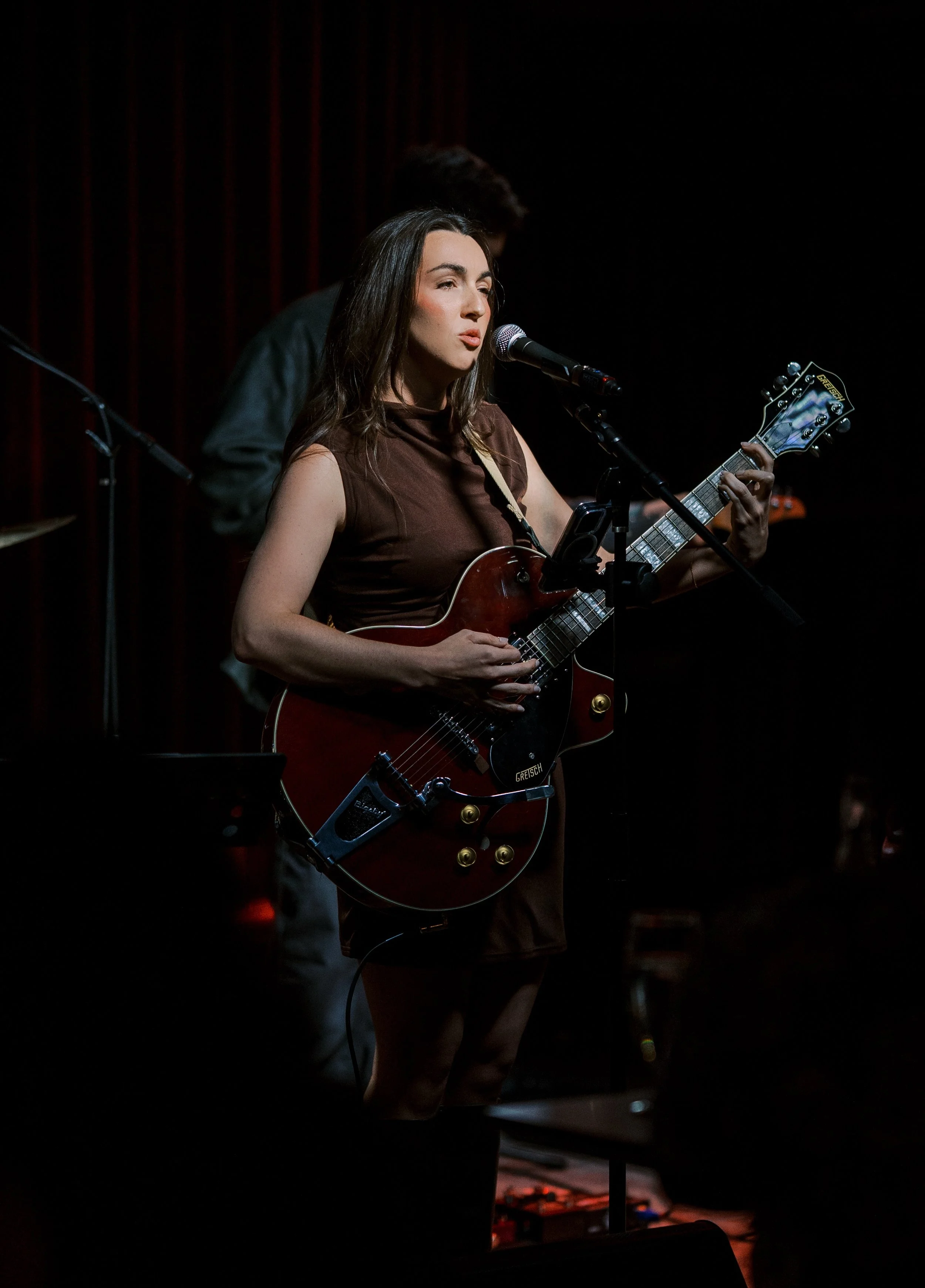 A woman singing into a microphone while playing a red electric guitar on a dark stage.