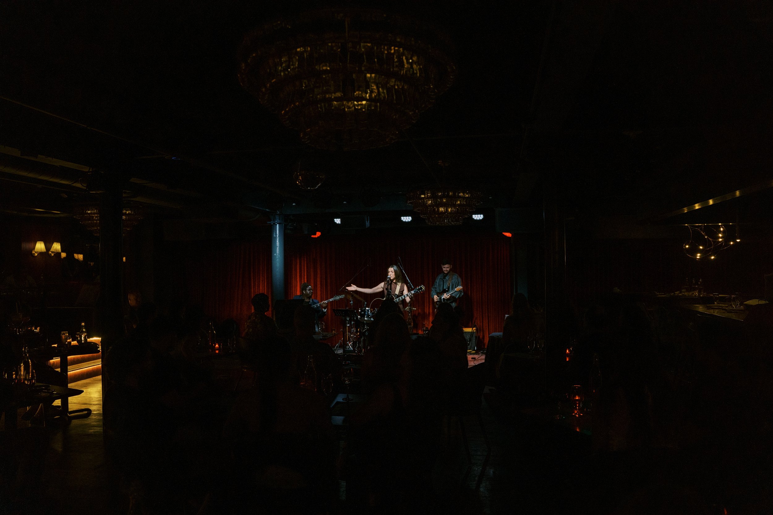 A musical performance on stage with musicians playing guitars and singing, illuminated by warm lighting in a dark venue with an audience watching.