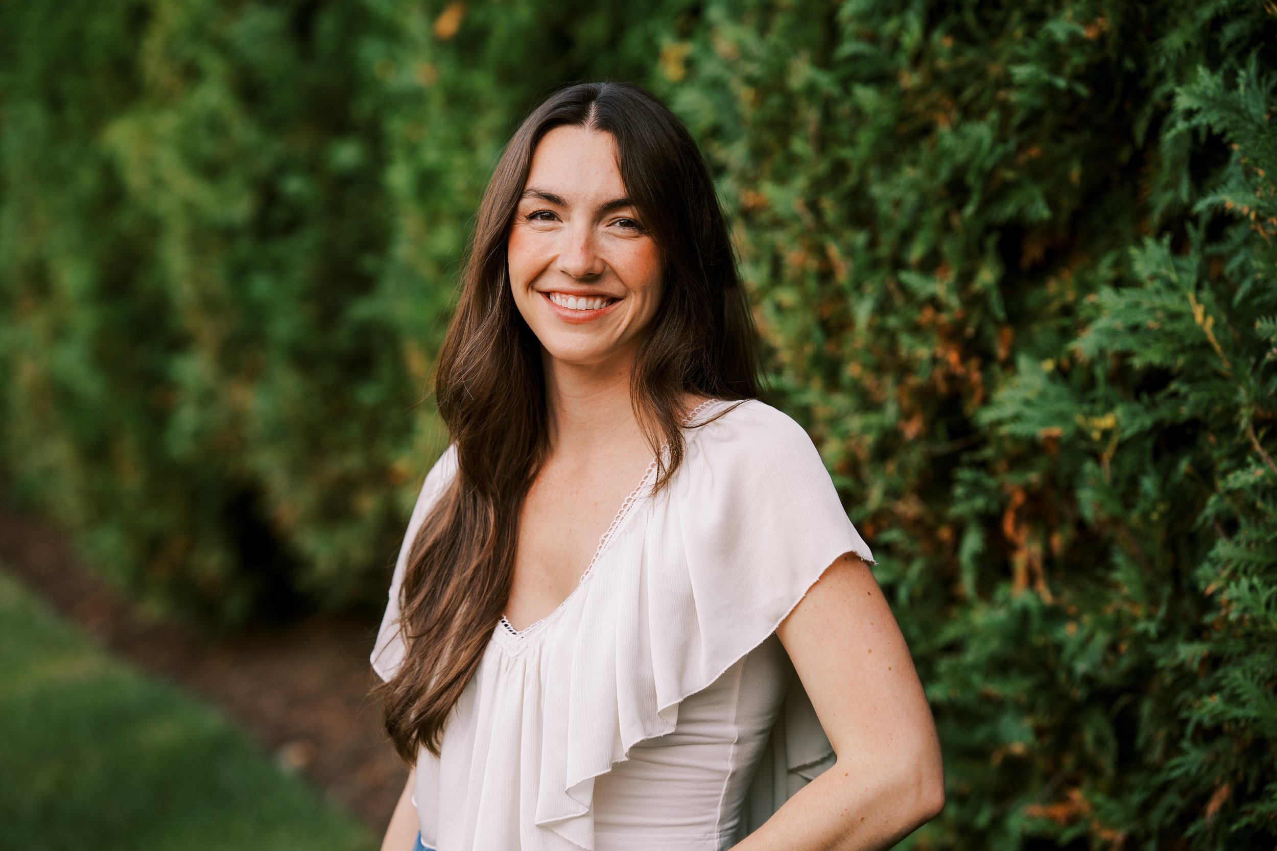 A woman with long brown hair smiling outdoors in front of green foliage.
