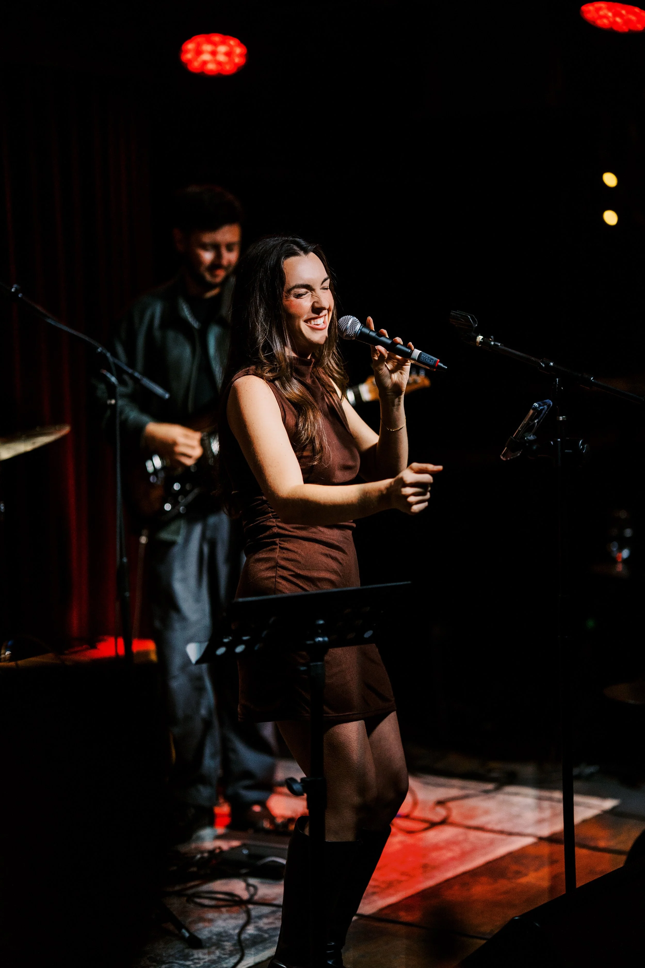 A woman holding a microphone singing on stage with a man playing guitar in the background, stage lighting with red and yellow spotlights, and music equipment on the floor.