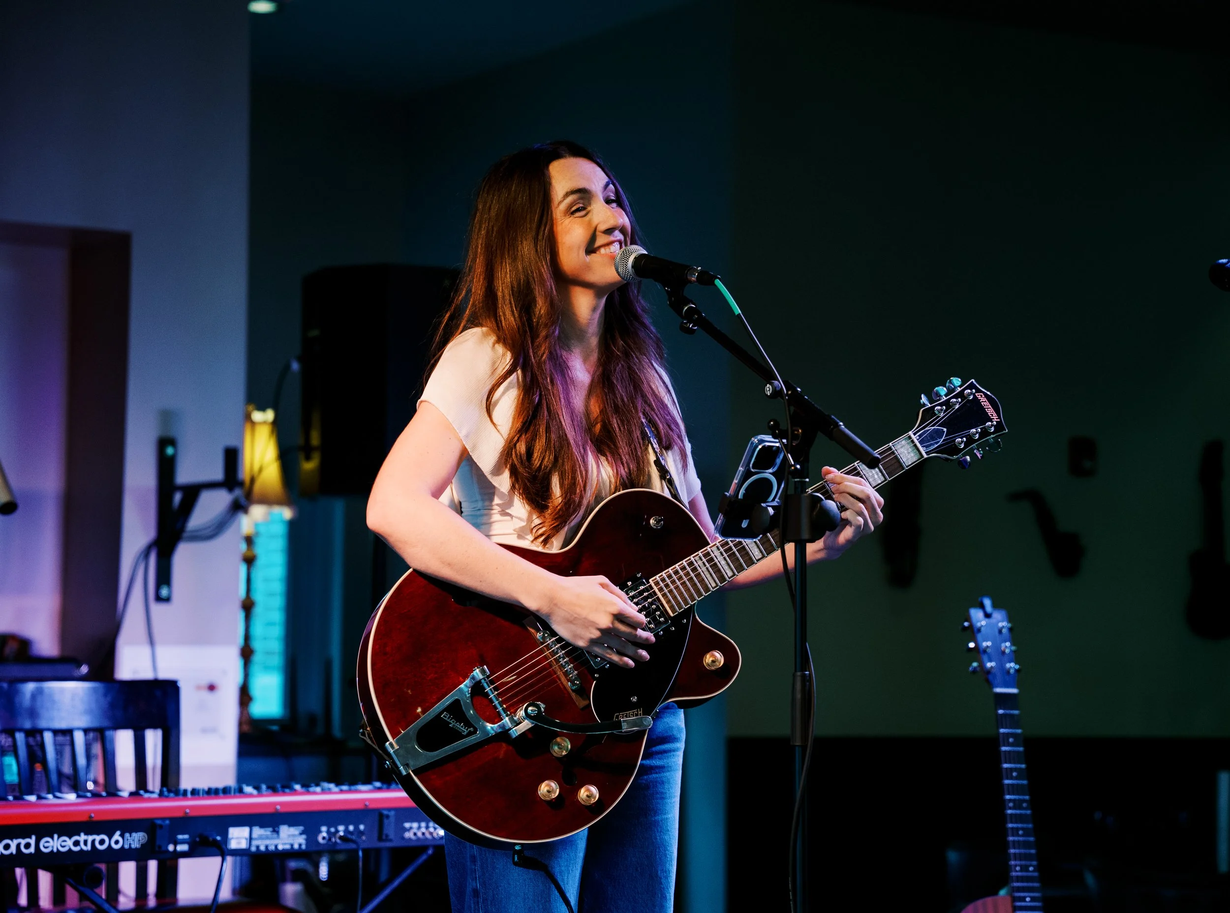 A woman with long brown hair smiling as she plays an electric guitar and sings into a microphone in a dimly lit music room with various instruments and equipment.