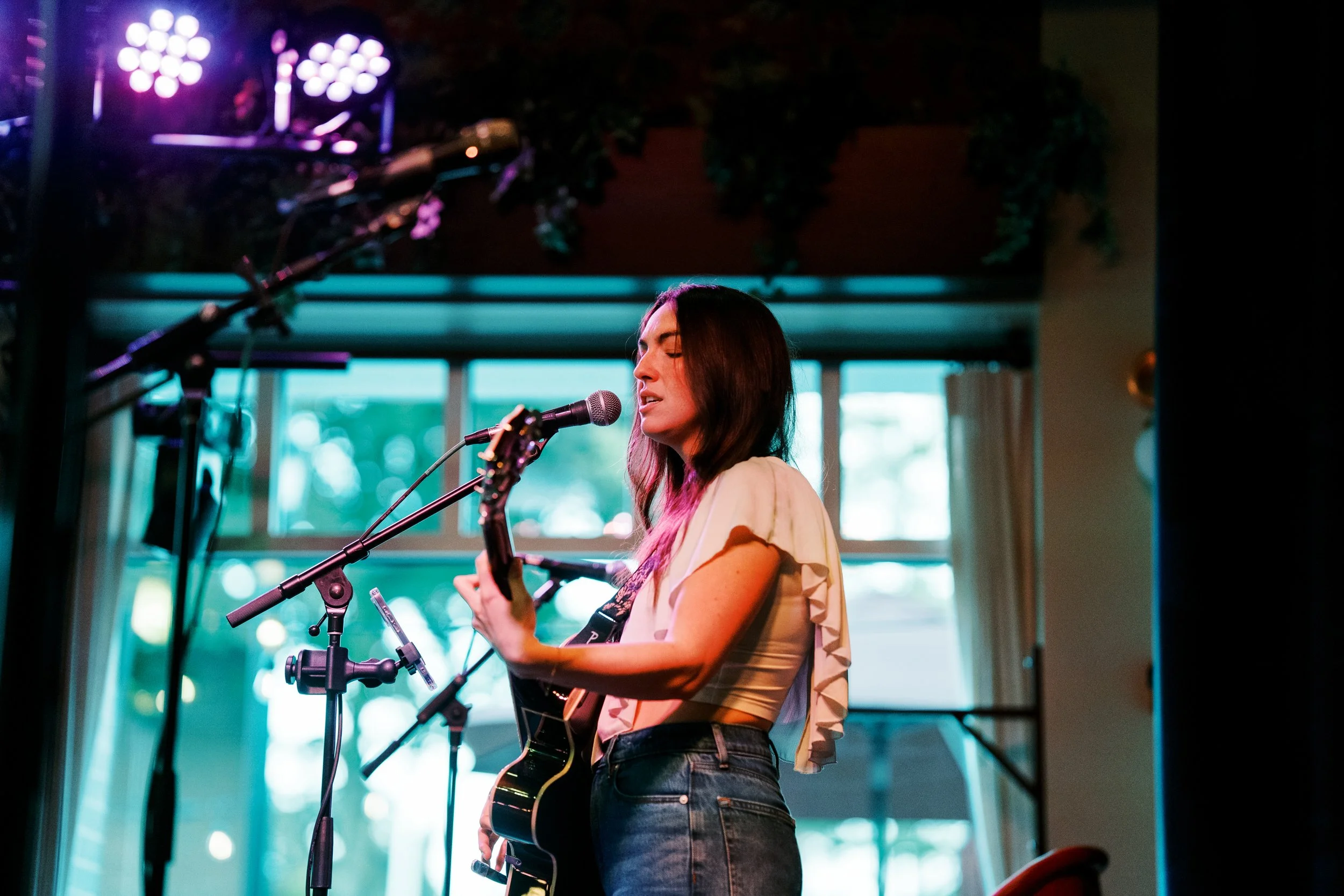 A woman with long brown hair, wearing a white top, is singing into a microphone while playing an acoustic guitar. She is in a room with large windows and decorative green plants behind her.