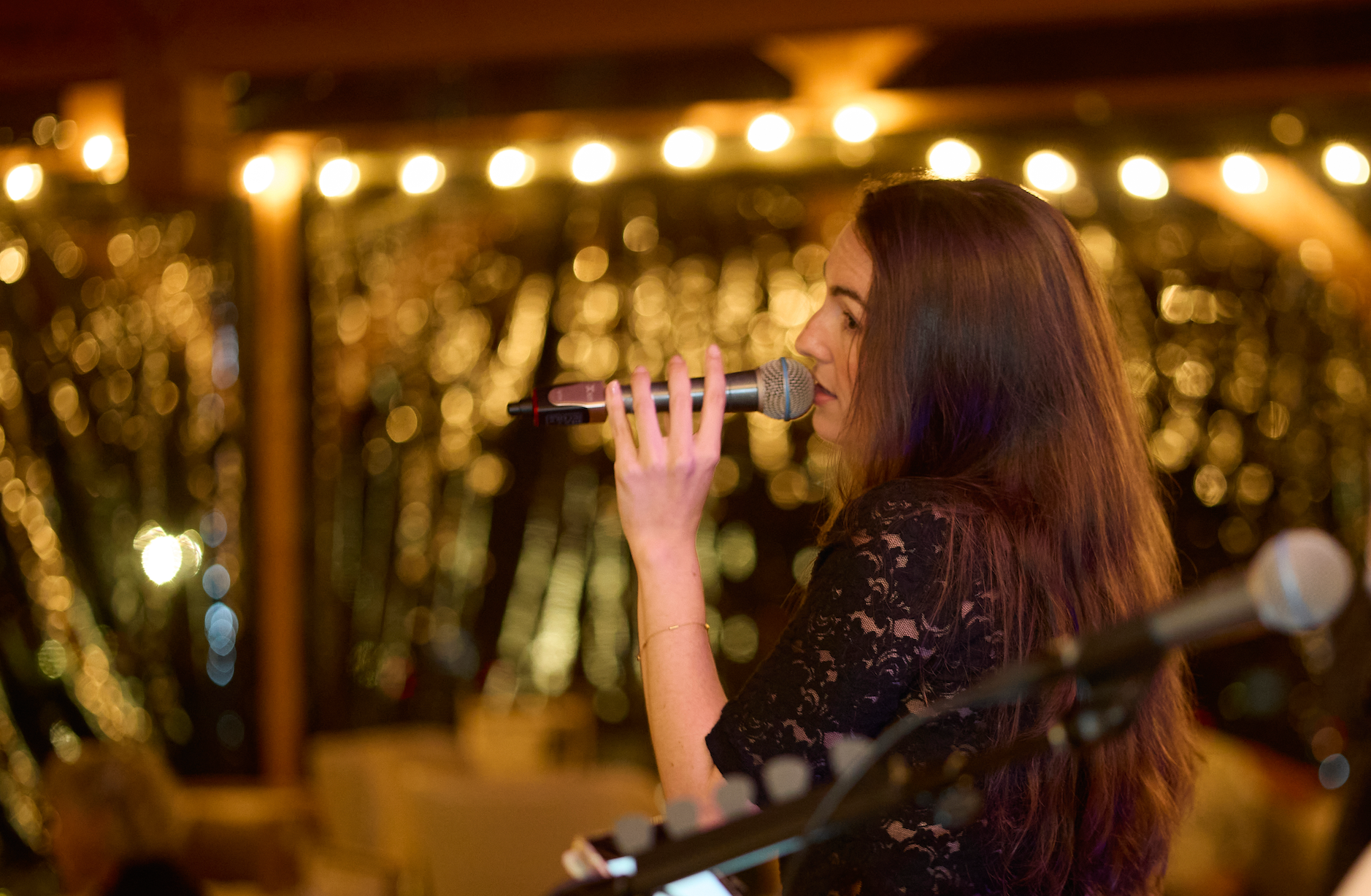 A woman with long brown hair singing into a microphone in a warmly lit setting with bokeh fairy lights in the background.