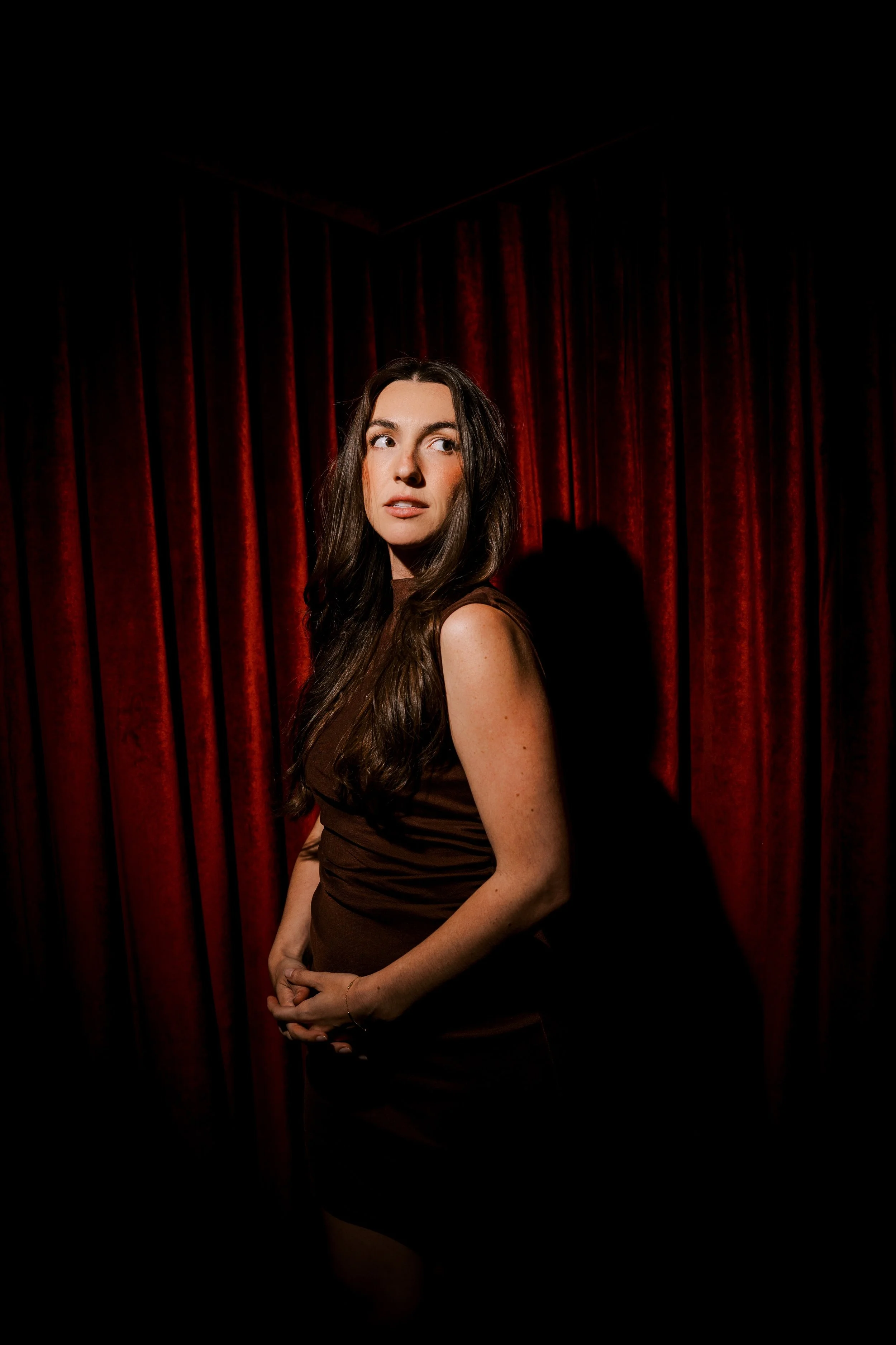 A woman with long brown hair, wearing a brown sleeveless dress, standing against red velvet curtains, looking to her left.