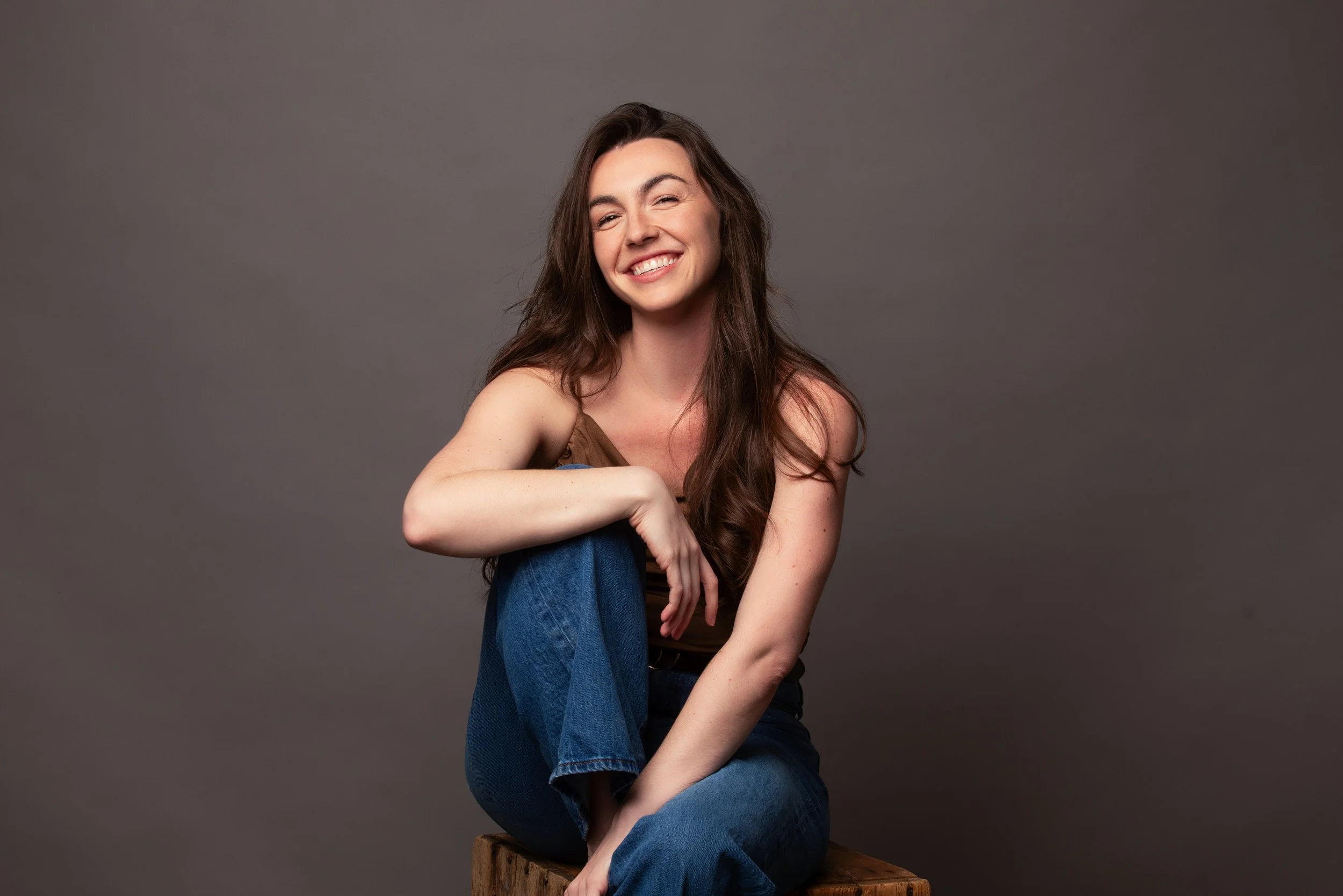 A woman with long brown hair, smiling, sitting on a wooden block against a gray background, wearing a brown tank top and blue jeans.