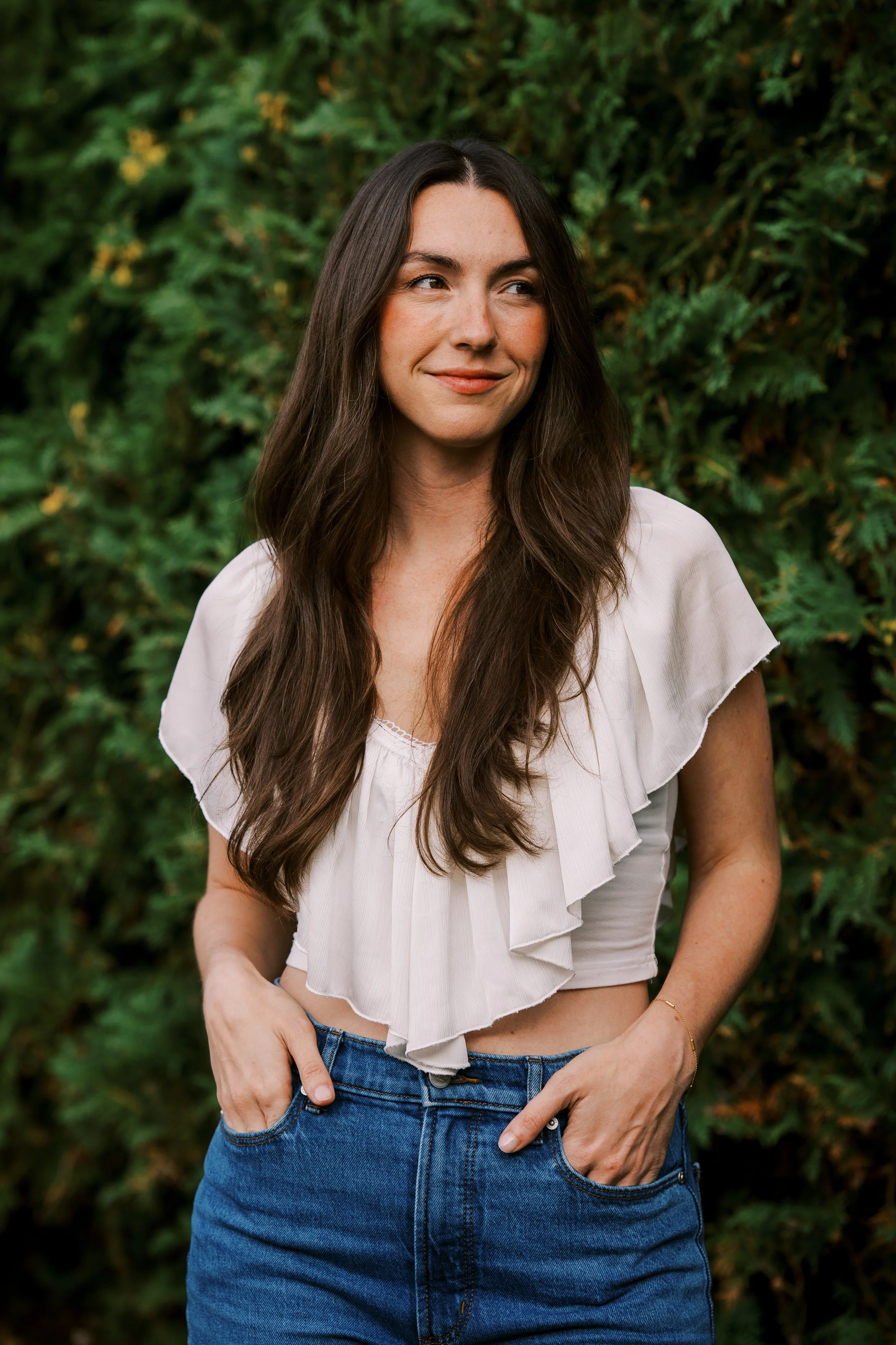 A woman with long brown hair, wearing a white ruffled top and blue jeans, standing outdoors in front of greenery, with her hands in her pockets, looking to the side and smiling softly.