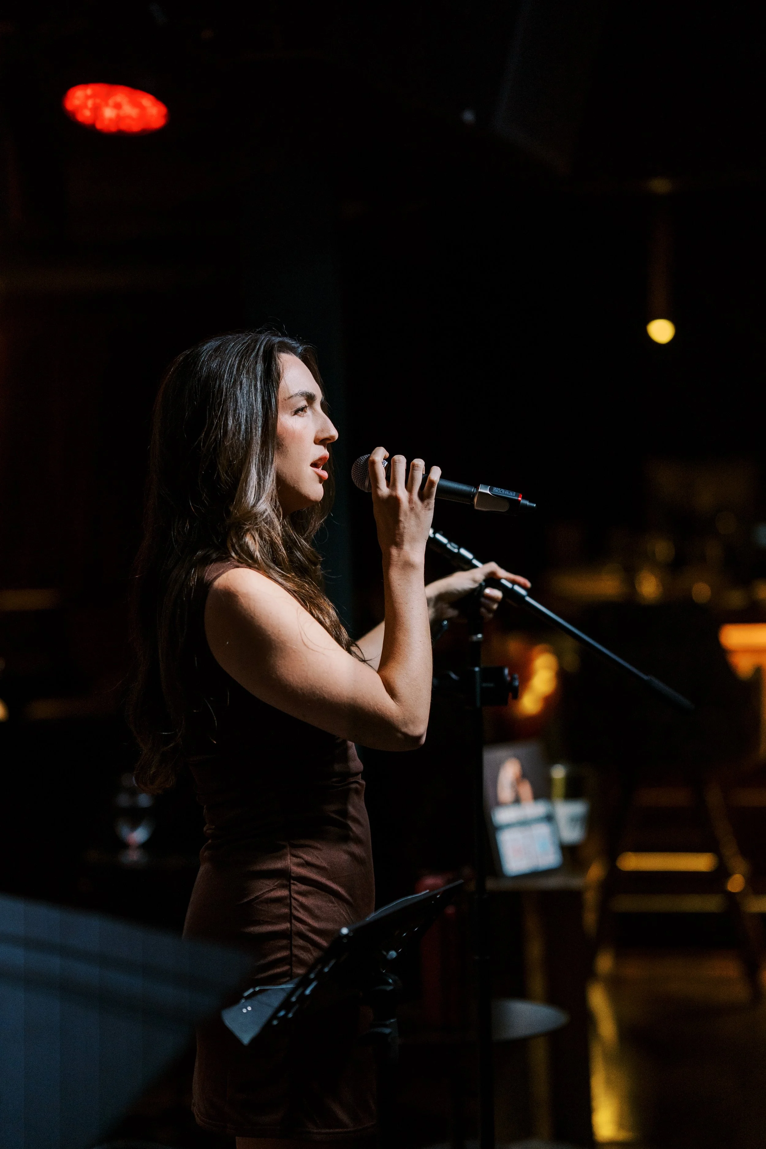 A woman with long wavy hair singing into a microphone on a dark stage.