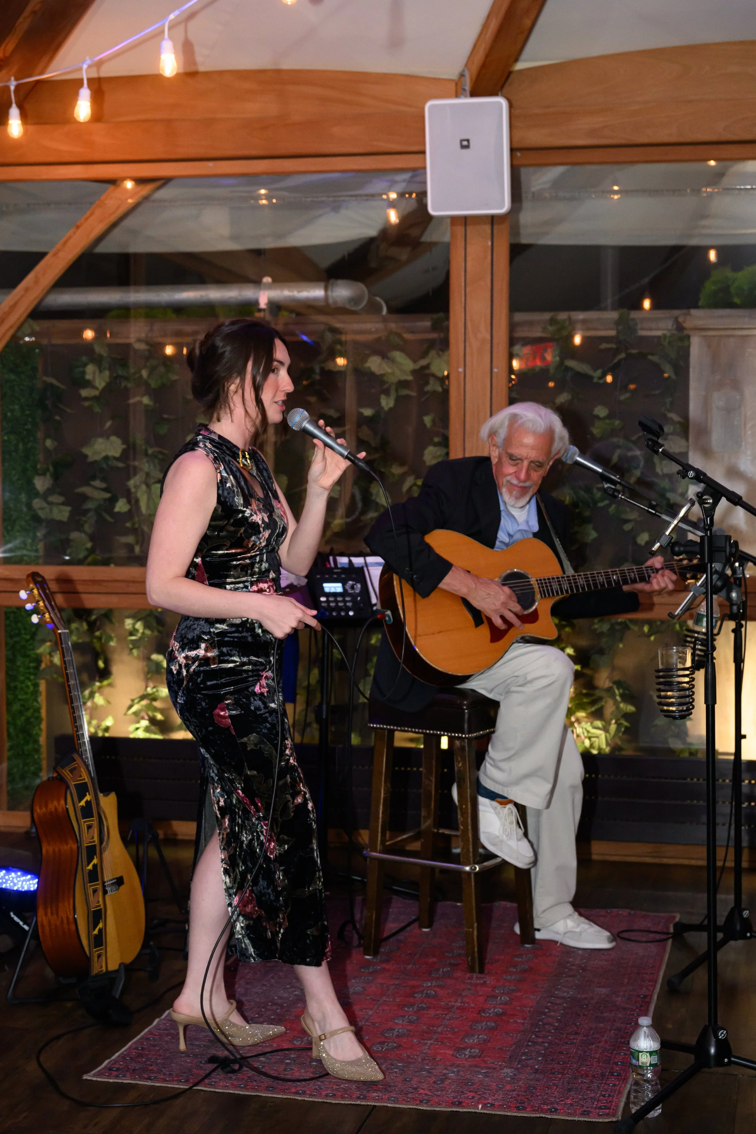 A woman singing into a microphone and a man playing an acoustic guitar, sitting on a stool, performing in a cozy, decorated venue with hanging string lights and green plants in the background.