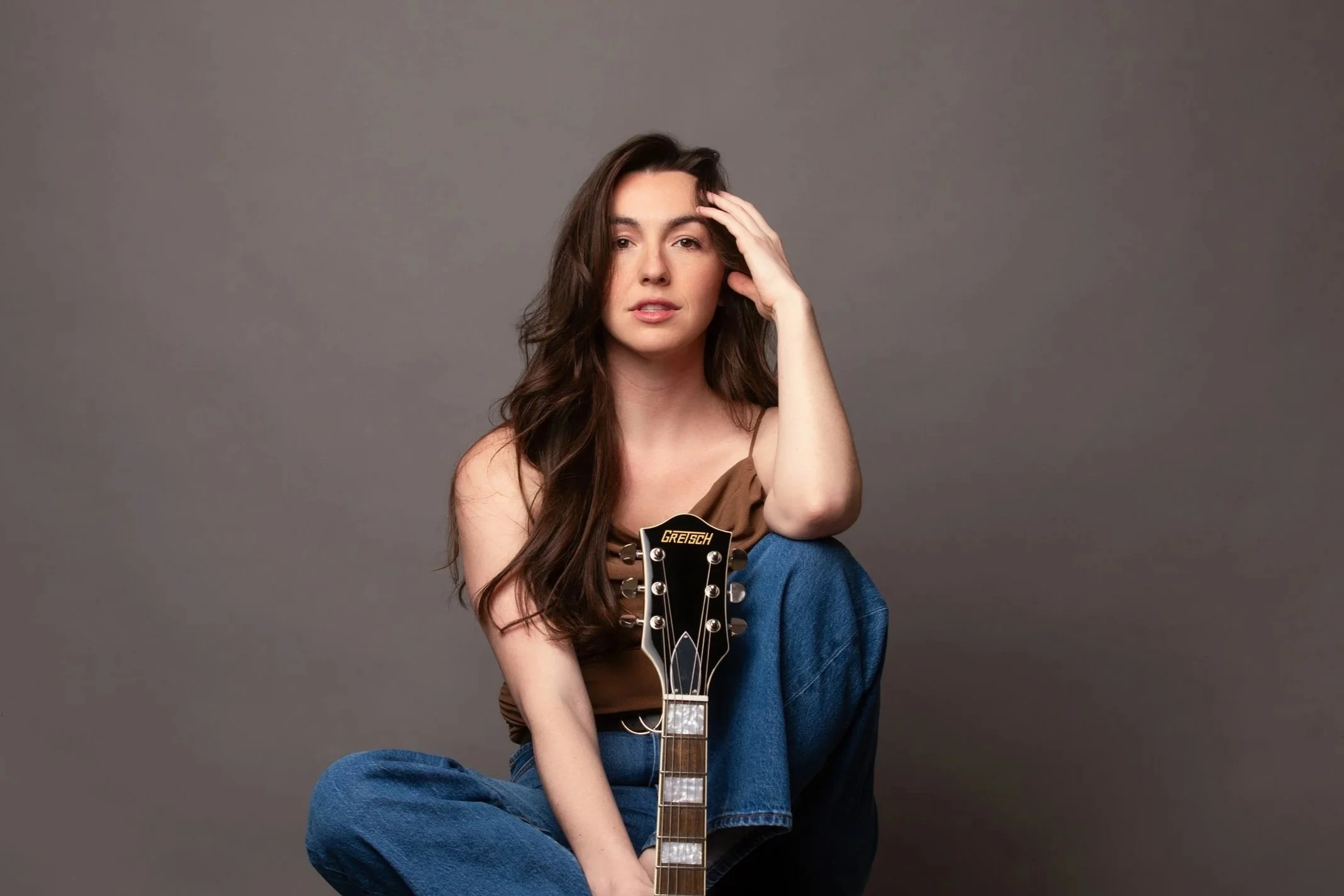 A woman sitting cross-legged on the floor holding an acoustic guitar, wearing a brown top and blue jeans, with a neutral gray background.