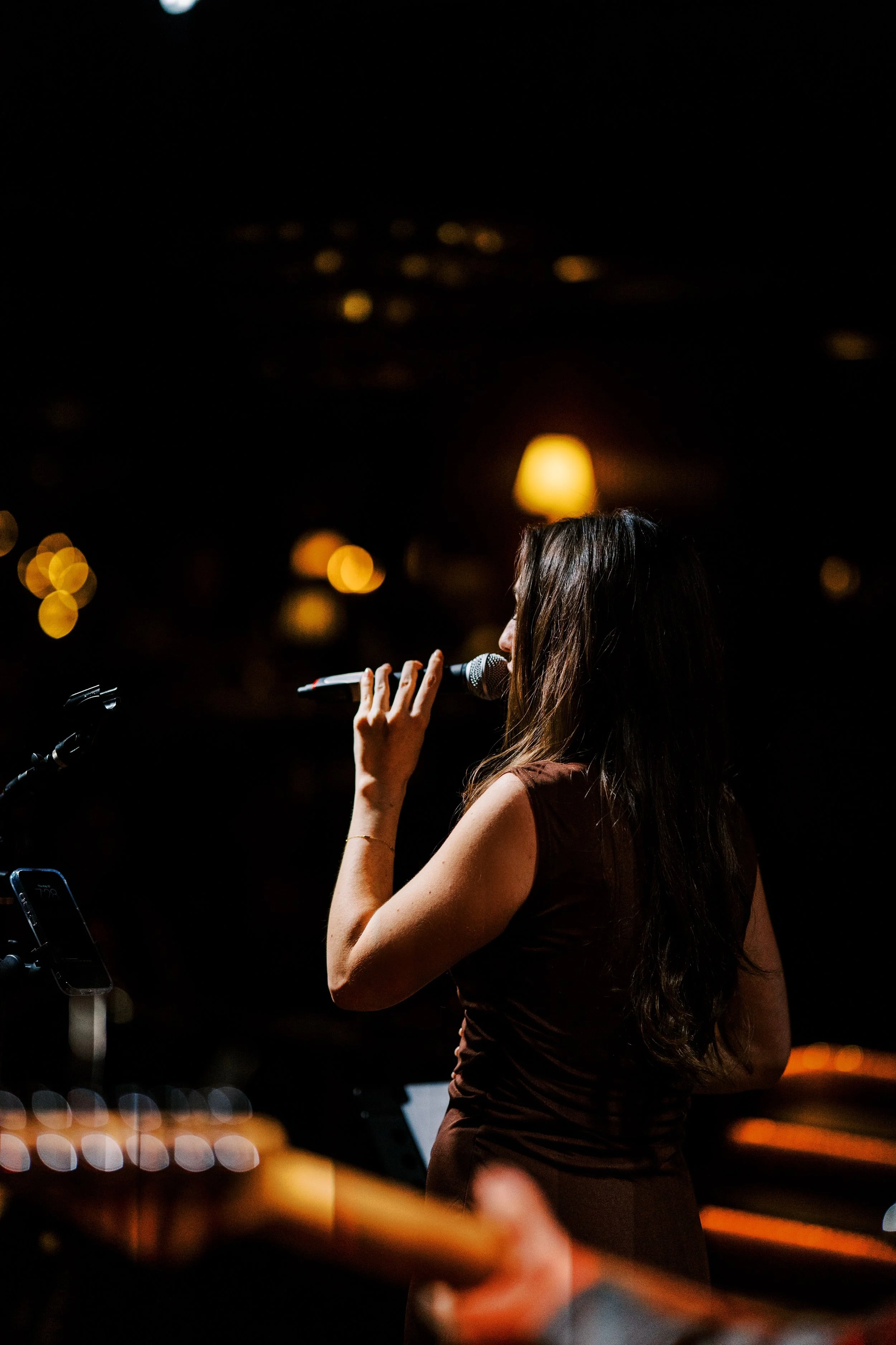 A woman with long hair singing into a microphone on stage at night, with blurred city lights in the background.