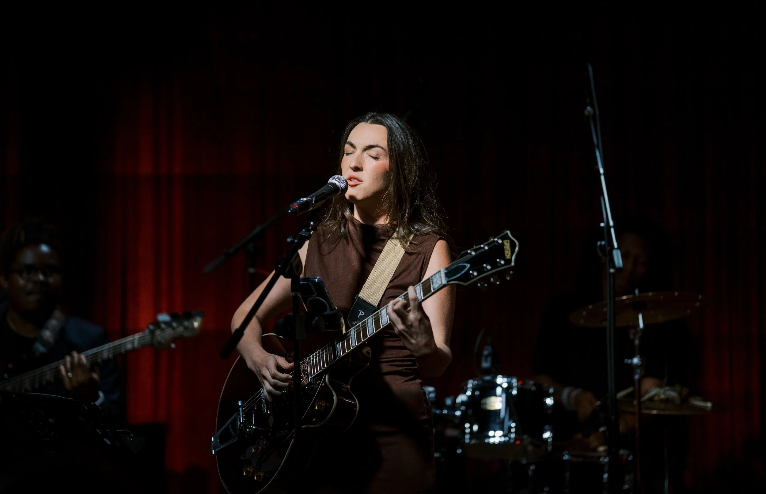 A woman with dark hair singing and playing an electric guitar on stage with closed eyes, accompanied by band members in the background.